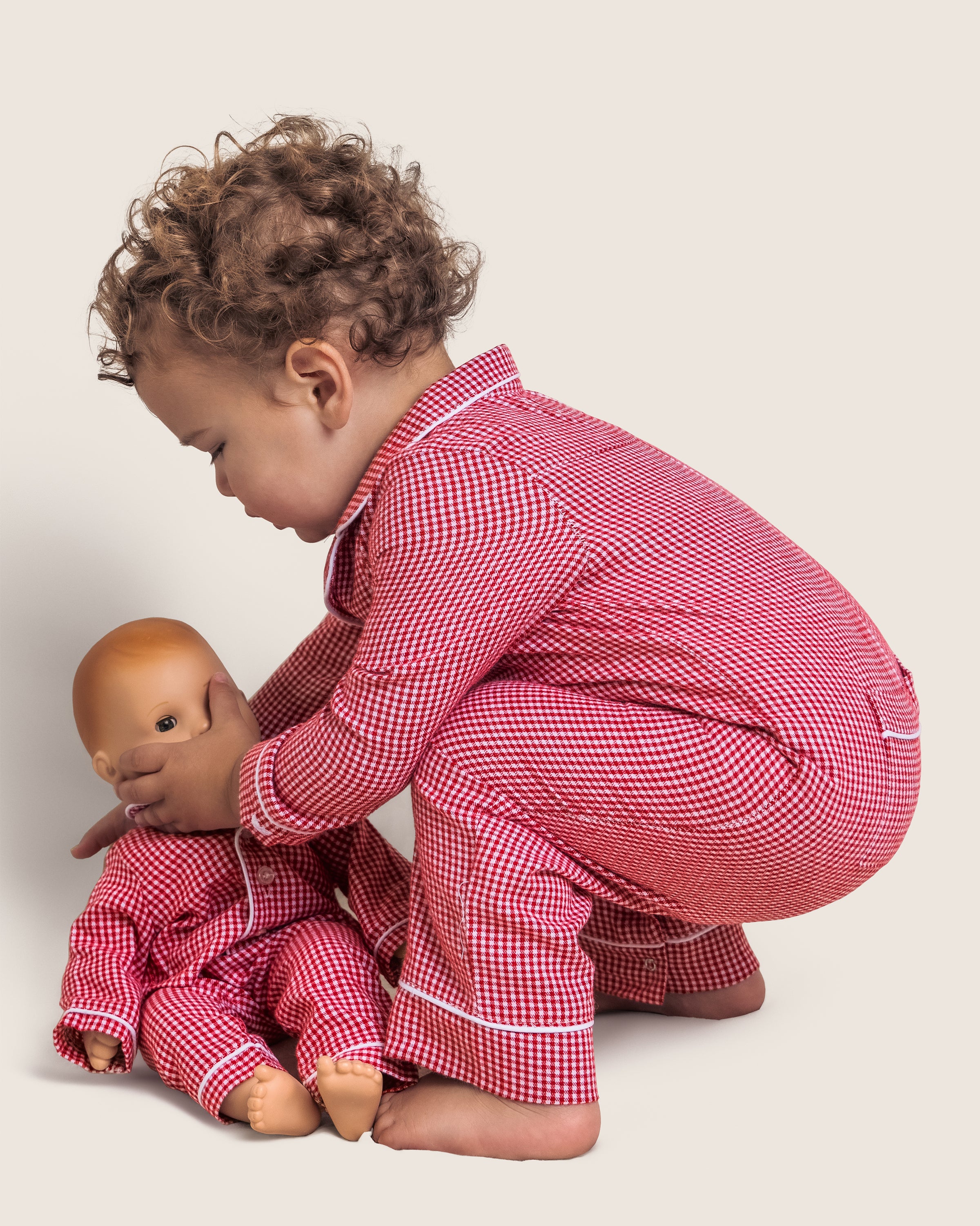 A toddler with curly hair wears Petite Plumes Babys Flannel Romper in Red Mini Gingham and plays with a matching doll, gently touching its face. They are set against a light, plain background that enhances the charming scene.