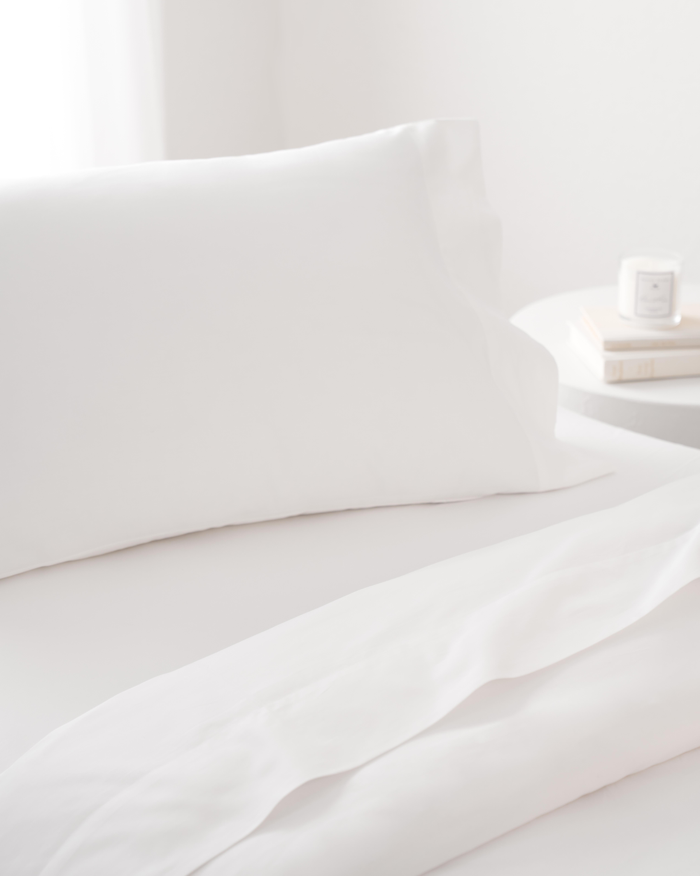 A neatly made bed showcases Petite Plumes Signature Luxe Sateen Bed Sheets in white, accompanied by crisp pillowcases. Beside the bed, a round white table holds books and a lit candle against a softly lit backdrop, evoking a five-star hotel ambiance.