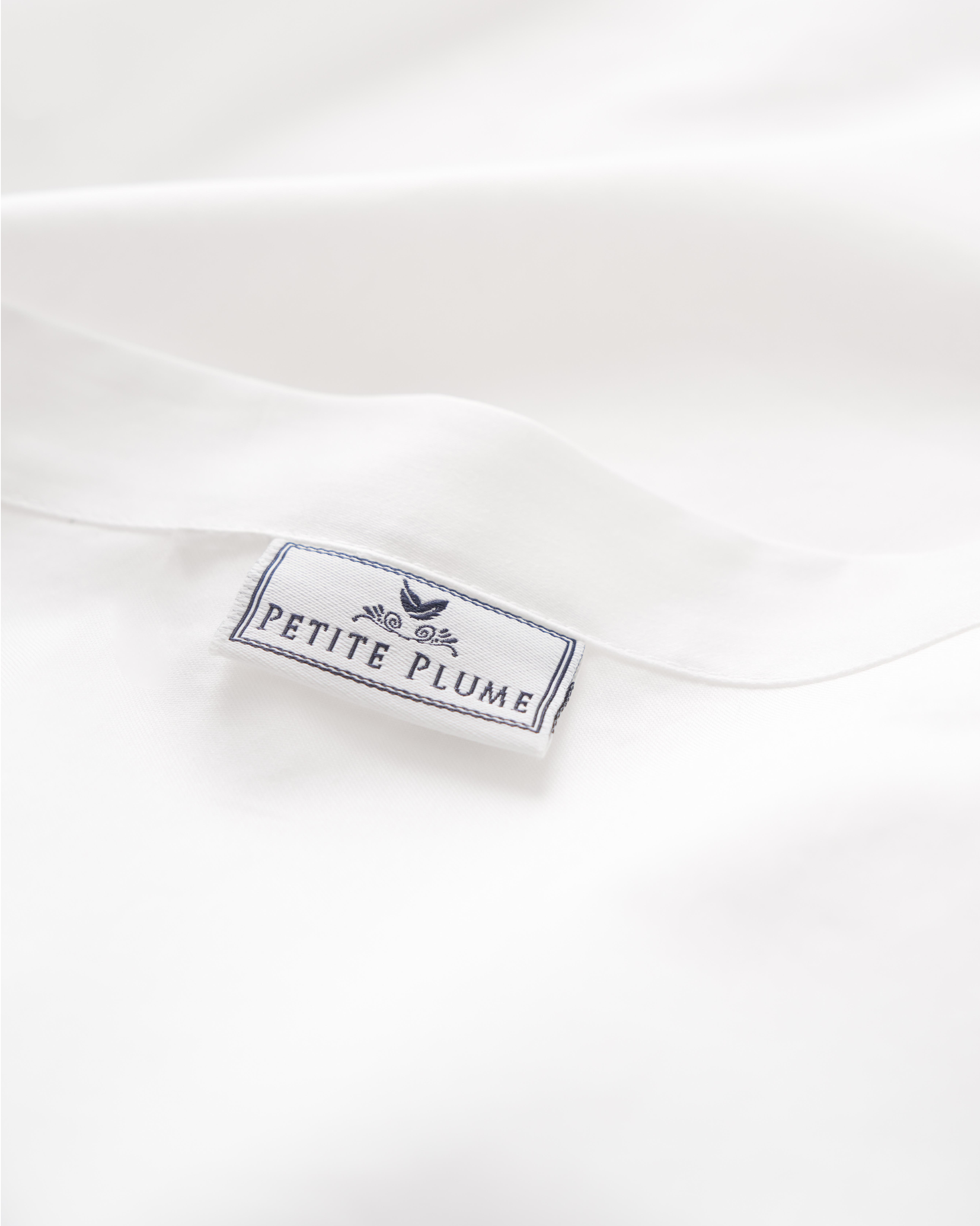 Close-up of a white sateen fabric label reading Petite Plume in blue, with decorative border and plume graphic, similar to those on Signature Luxe Sateen Bed Sheets by Petite Plume from luxury hotels.