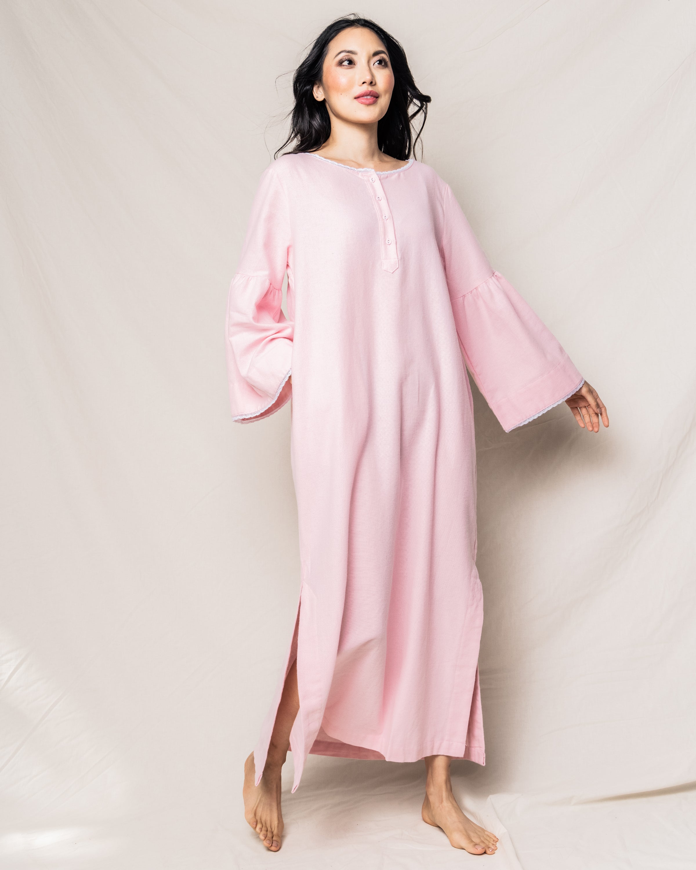 A woman poses barefoot against a light background in a Petite Plume Womens Flannel Seraphine Nightgown in Pink, featuring bell sleeves, button details on the front, and a slit near the hem. Her dark hair falls naturally.