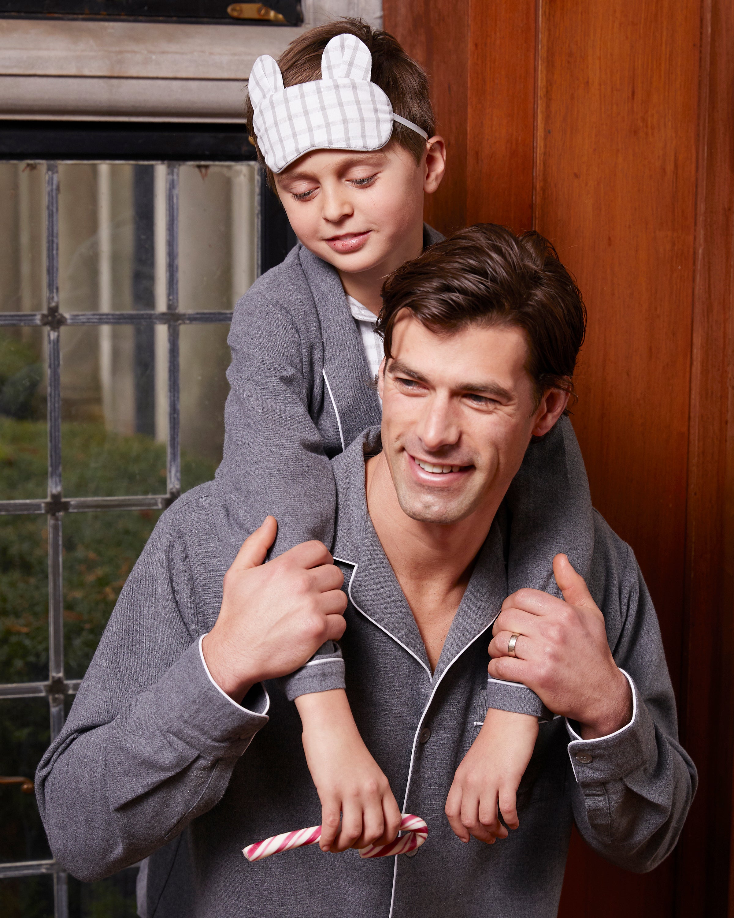 Inside a room, a man carries a young boy on his shoulders. Both wear matching Mens Flannel Pajama Sets in Grey by Petite Plume. The boy sports an adorable checkered sleep mask with bunny ears and holds a candy cane, their expressions joyfully relaxed.