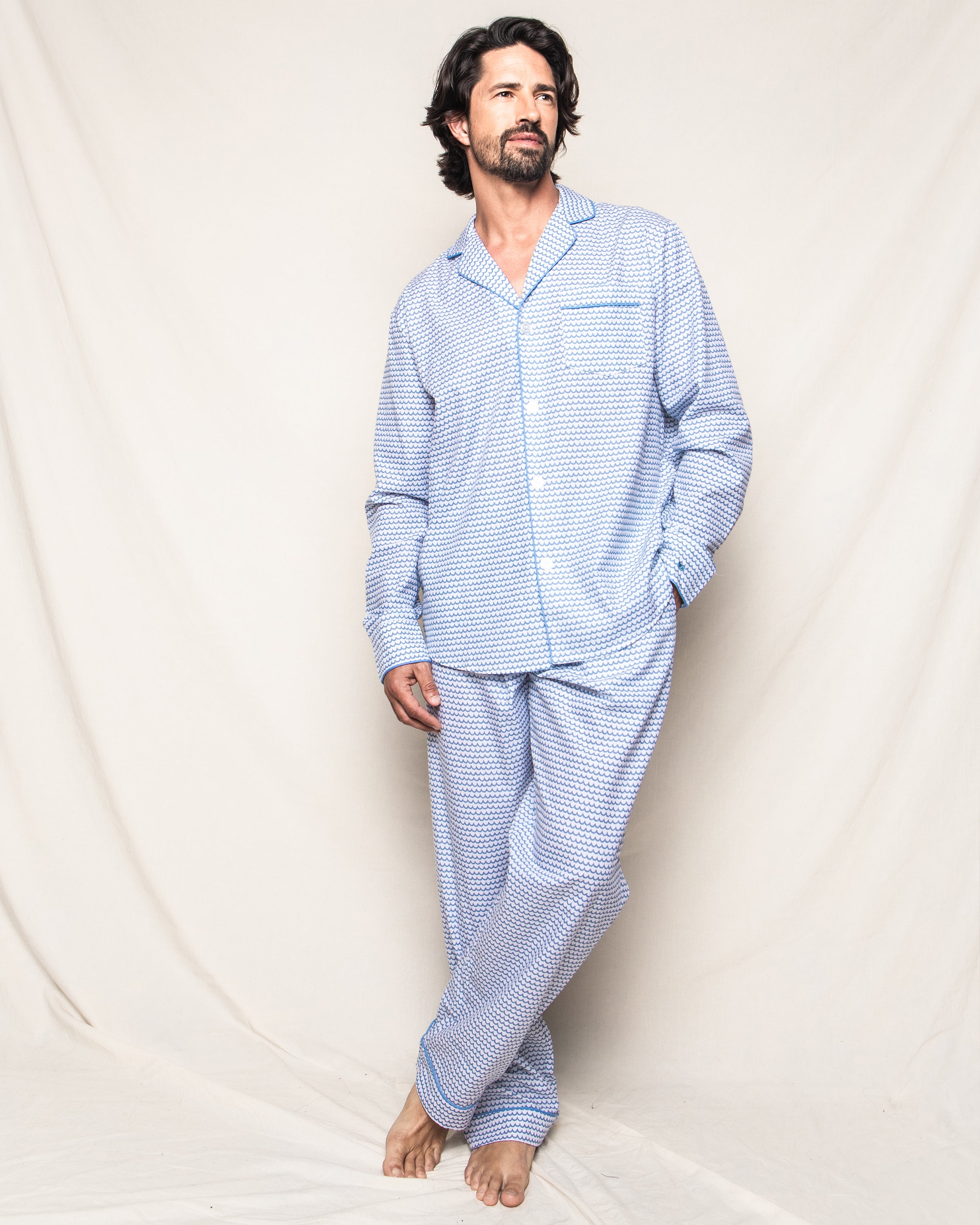 A dark-haired, bearded man models the Mens Twill Pajama Set in La Mer by Petite Plume, standing barefoot against a draped background with a relaxed look, showcasing luxurious sleepwear style.