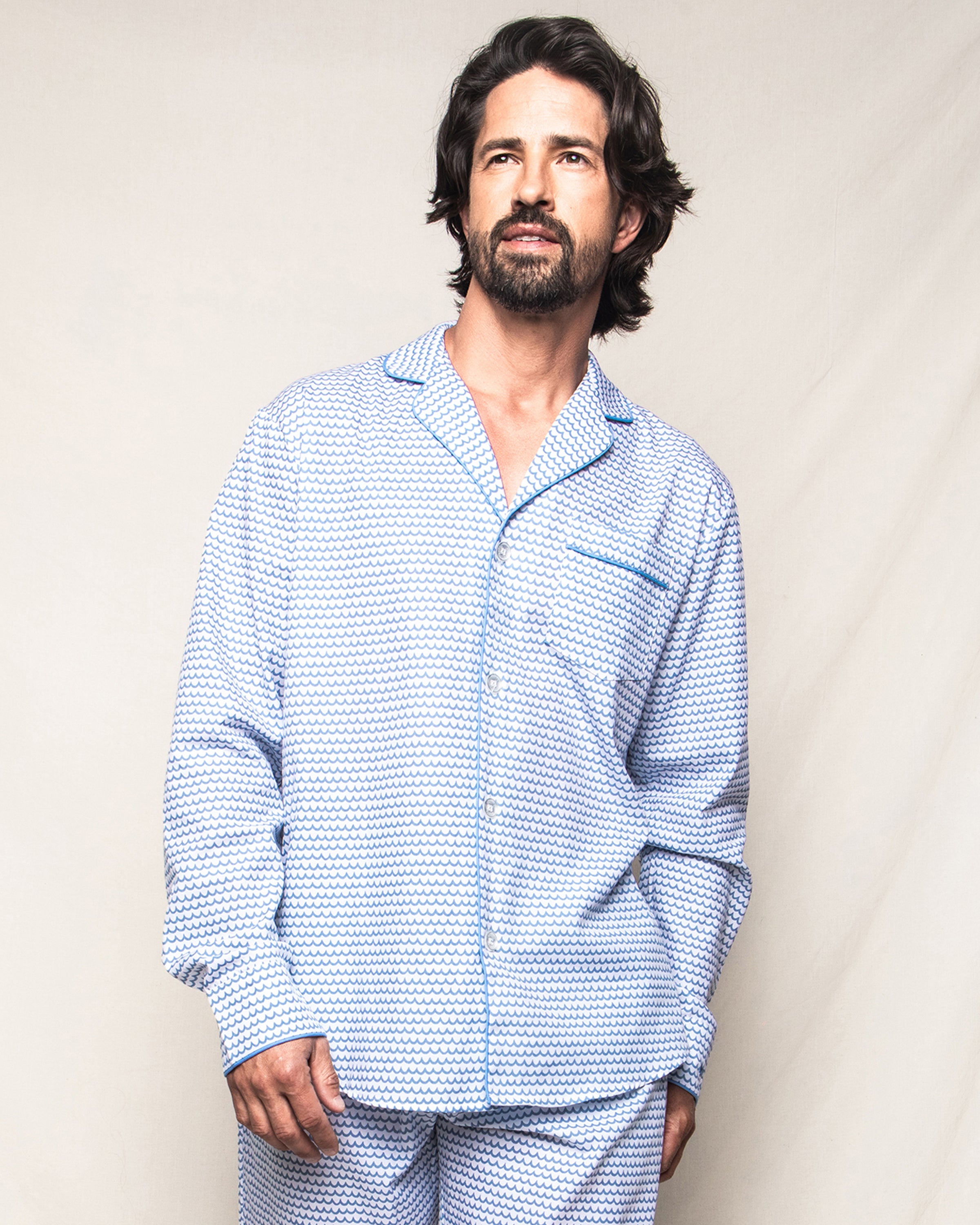 A man with shoulder-length hair and a beard stands against a plain light background, gazing slightly upward. Hes wearing the Petite Plume Mens Twill Pajama Set in La Mer, crafted from fine cotton in matching light blue and white patterns, epitomizing luxurious sleepwear.