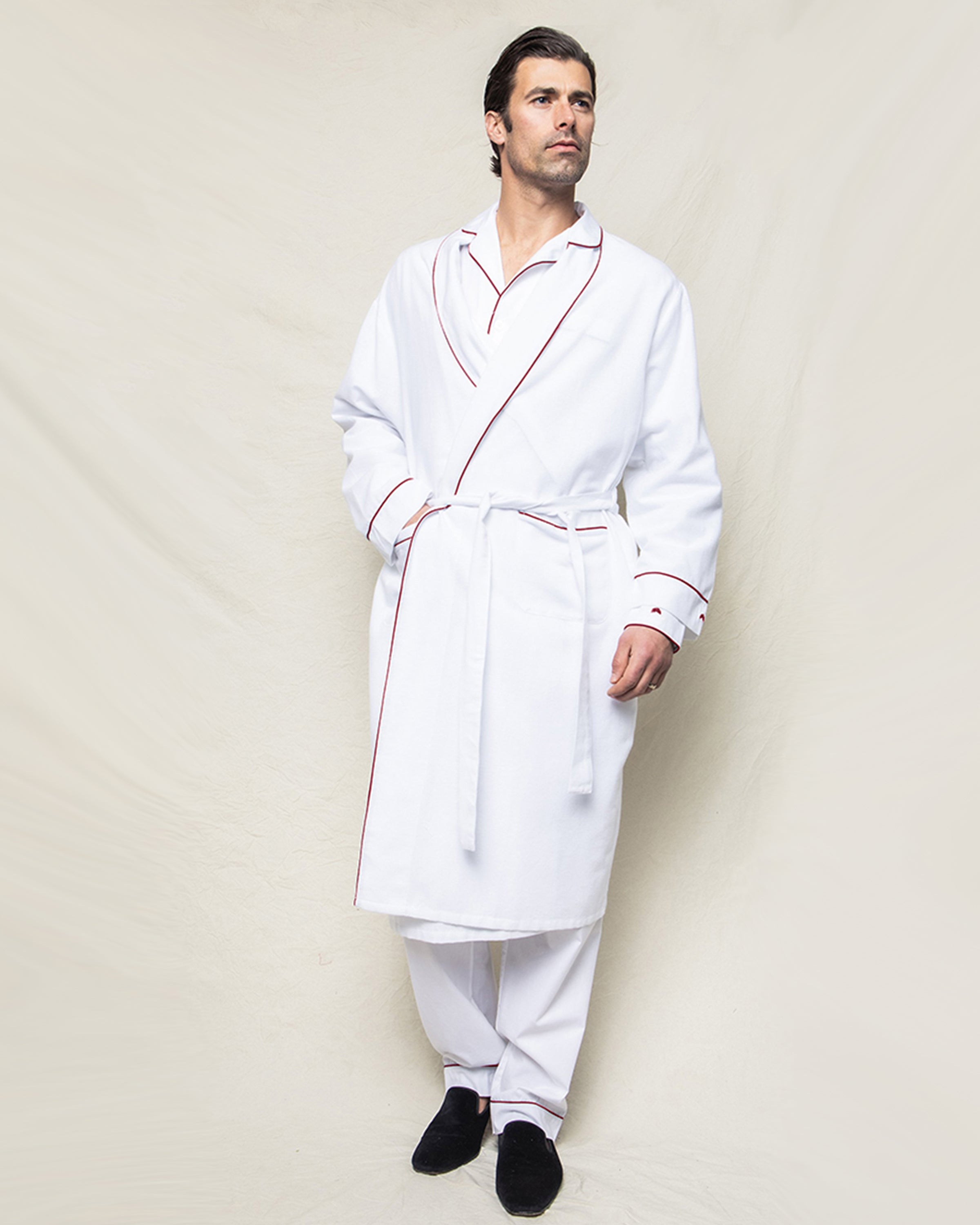 A person models Petite Plumes Mens Flannel Robe in White with Red Piping, against a neutral backdrop. With its elegant waist belt, its paired stylishly with white pants and dark slippers, creating a sophisticated look.