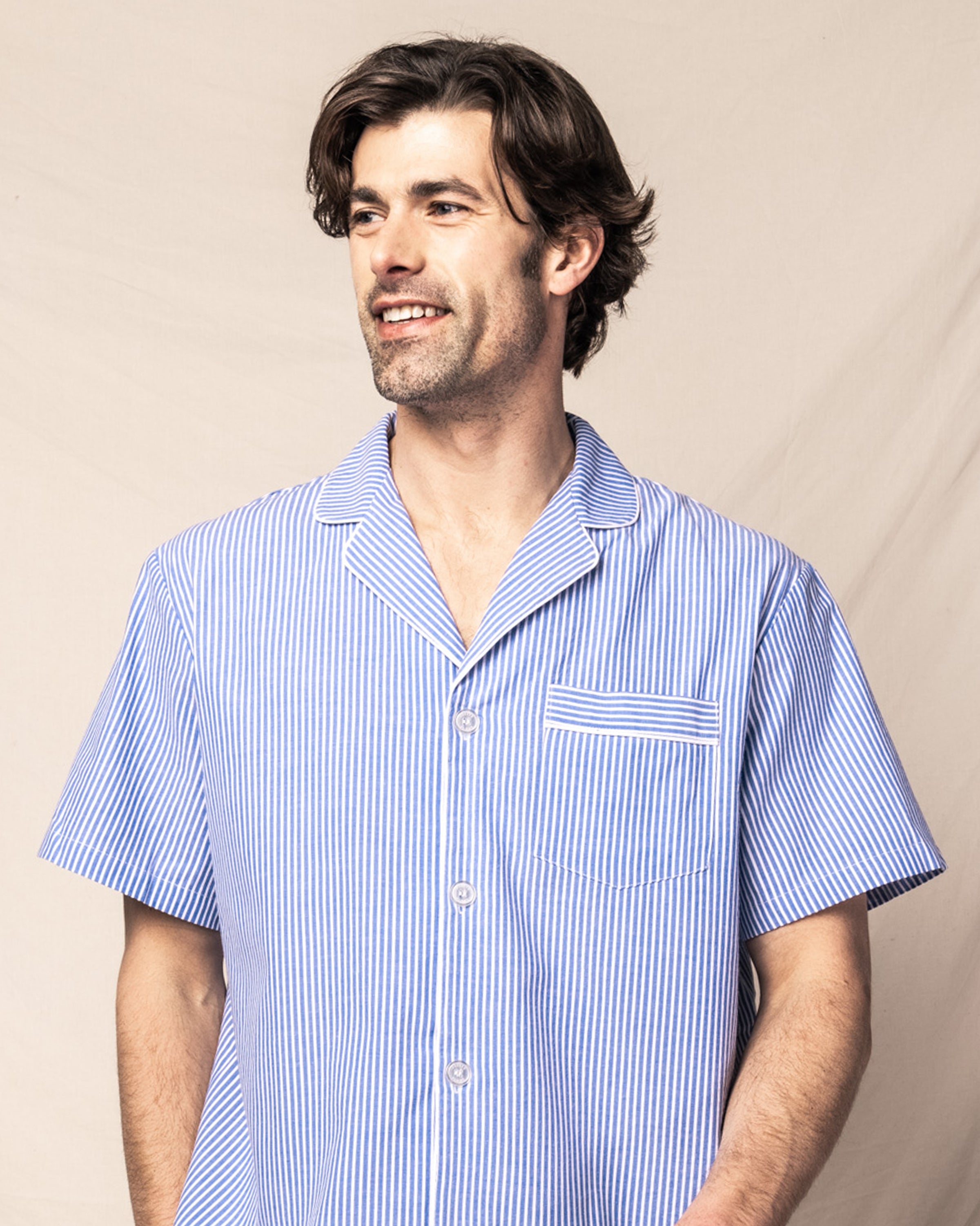 A dark-haired man smiles warmly, wearing Petite Plumes Mens Twill Pajama Short Set in French Blue Seersucker, made from 100% cotton with a left pocket. The light blue and white striped short-sleeve shirt exudes the comfort of luxury sleepwear against a plain backdrop.