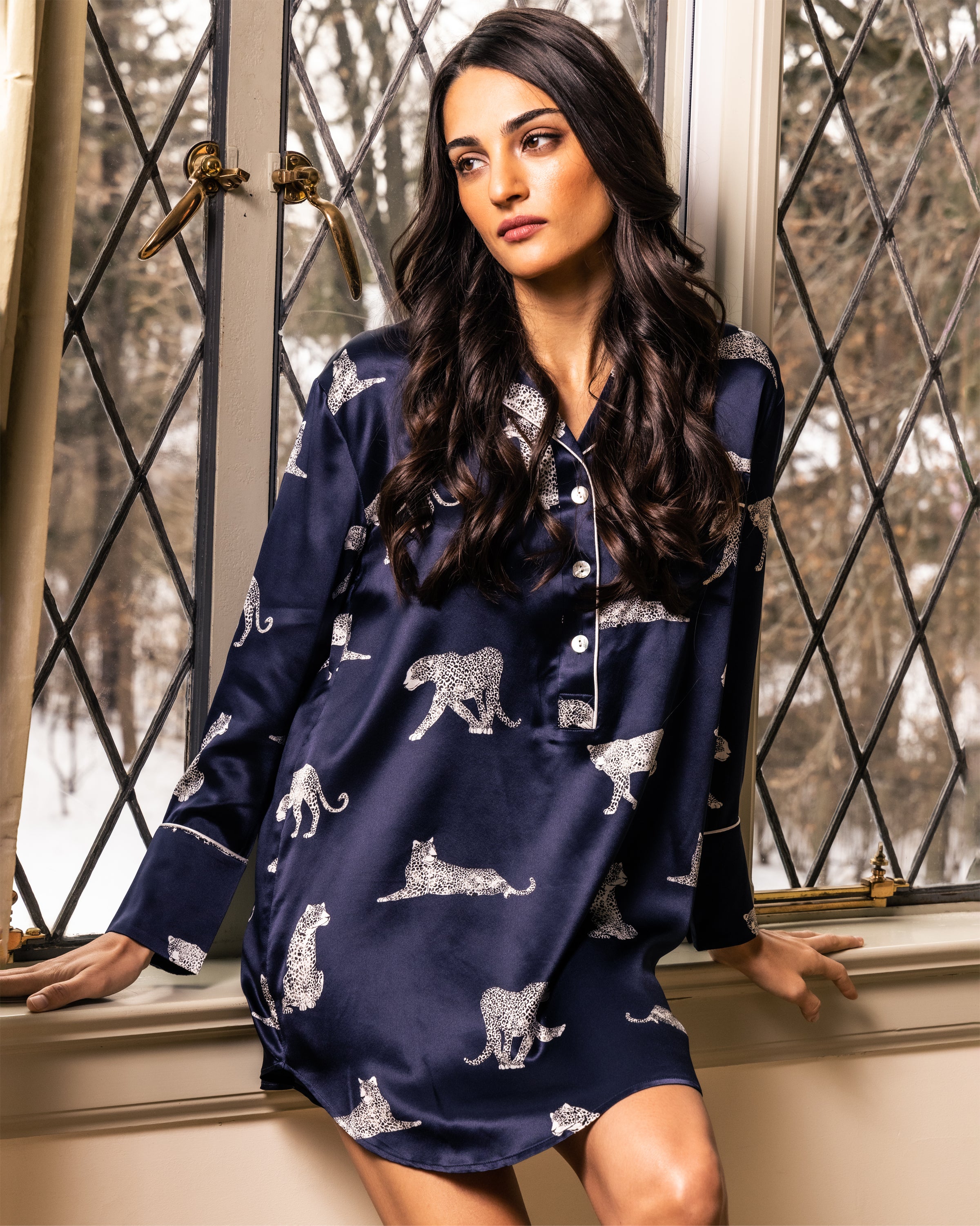 A person with long dark hair sits on a windowsill, wearing Petite Plumes Womens Silk Nightshirt in Panthère de Nuit. The windows diamond-shaped panes reveal a snowy scene, crafting the perfect backdrop for relaxation and style.