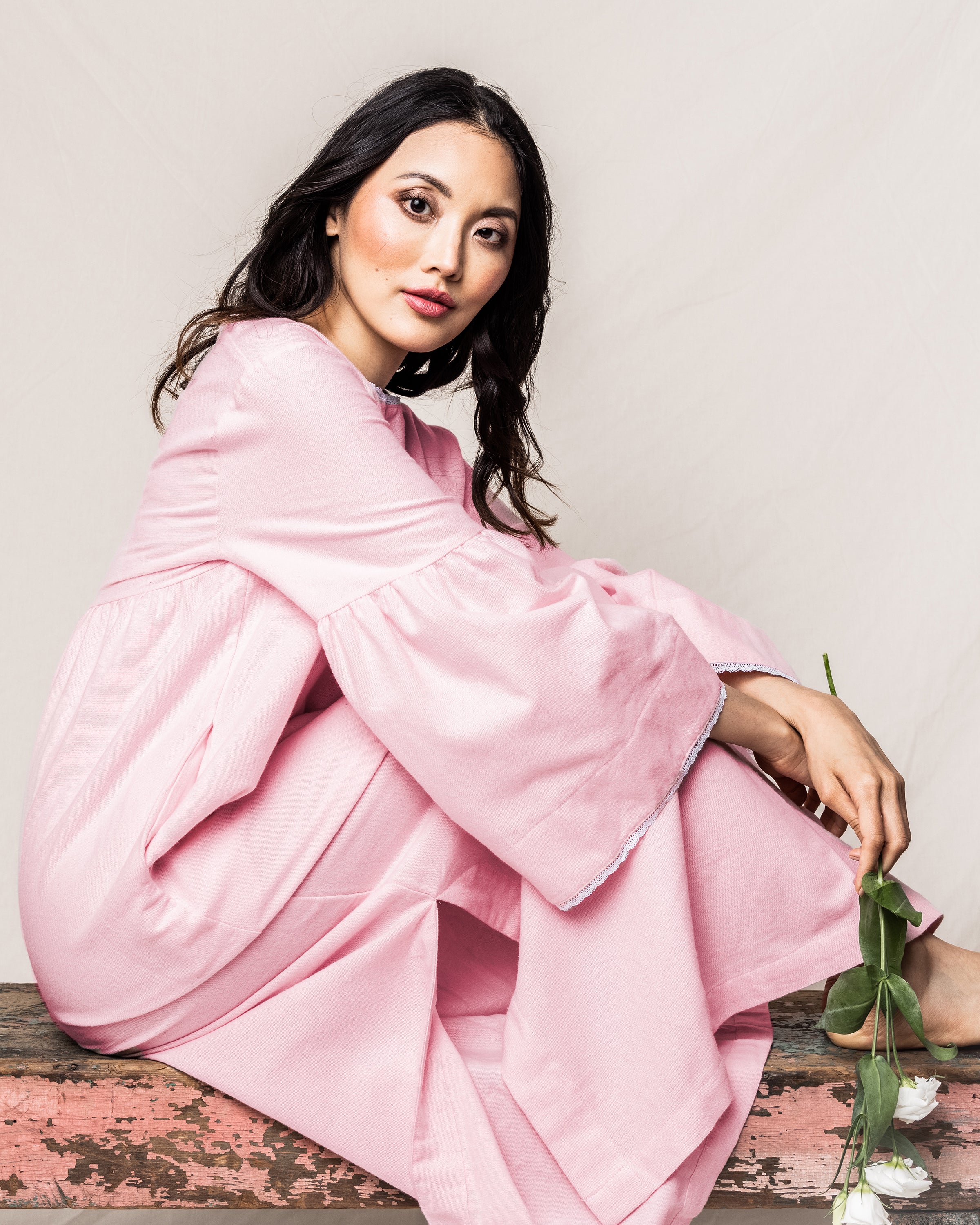 A person with long dark hair sits on a wooden bench wearing the Womens Flannel Seraphine Nightgown in Pink by Petite Plume. They hold a white flower, gazing softly at the camera against a plain, light-colored background that enhances the serene elegance of the scene.