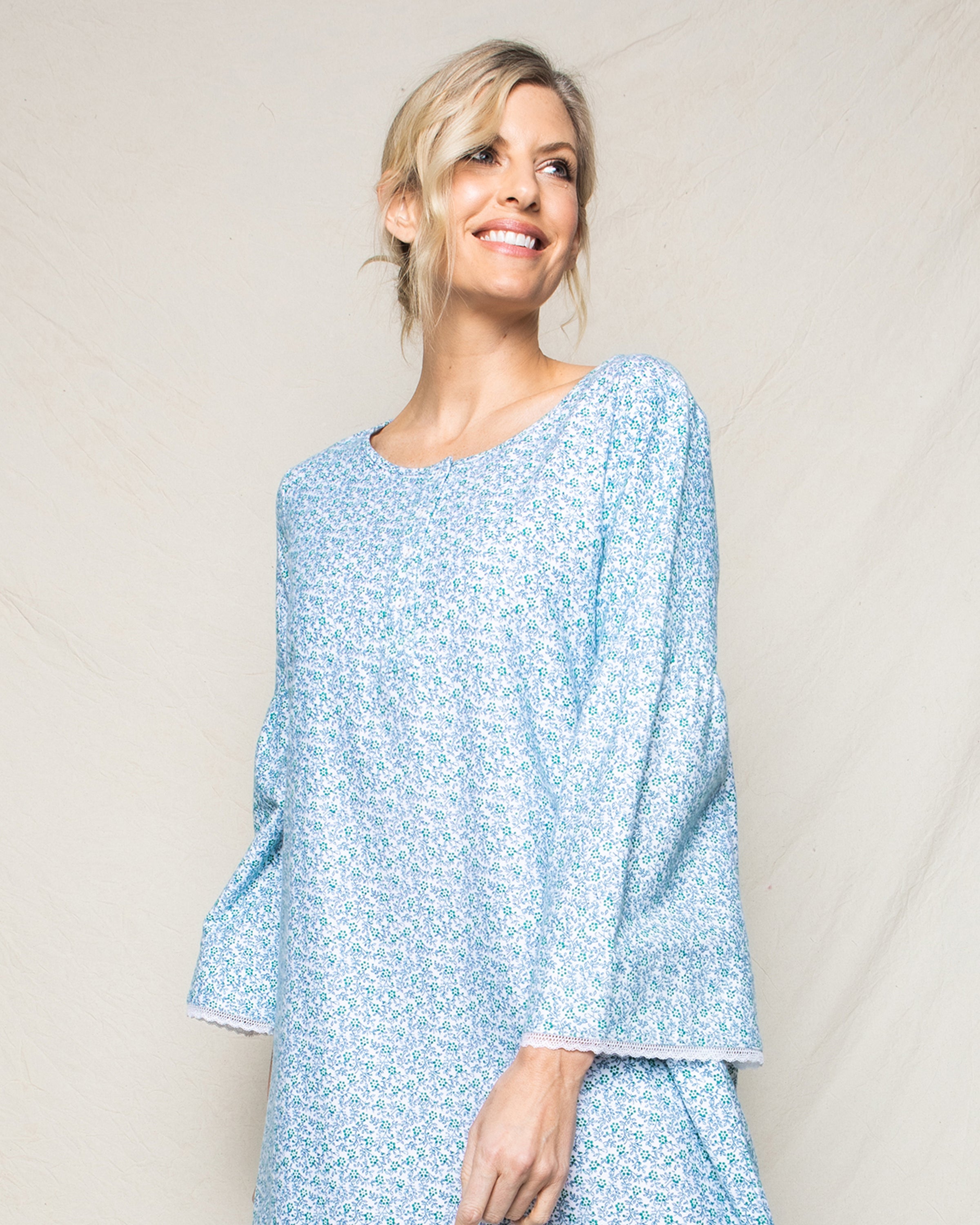 A blonde woman stands against a beige background, smiling and glancing sideways in a Petite Plume Womens Flannel Seraphine Nightgown featuring the Stafford Floral print in light blue.