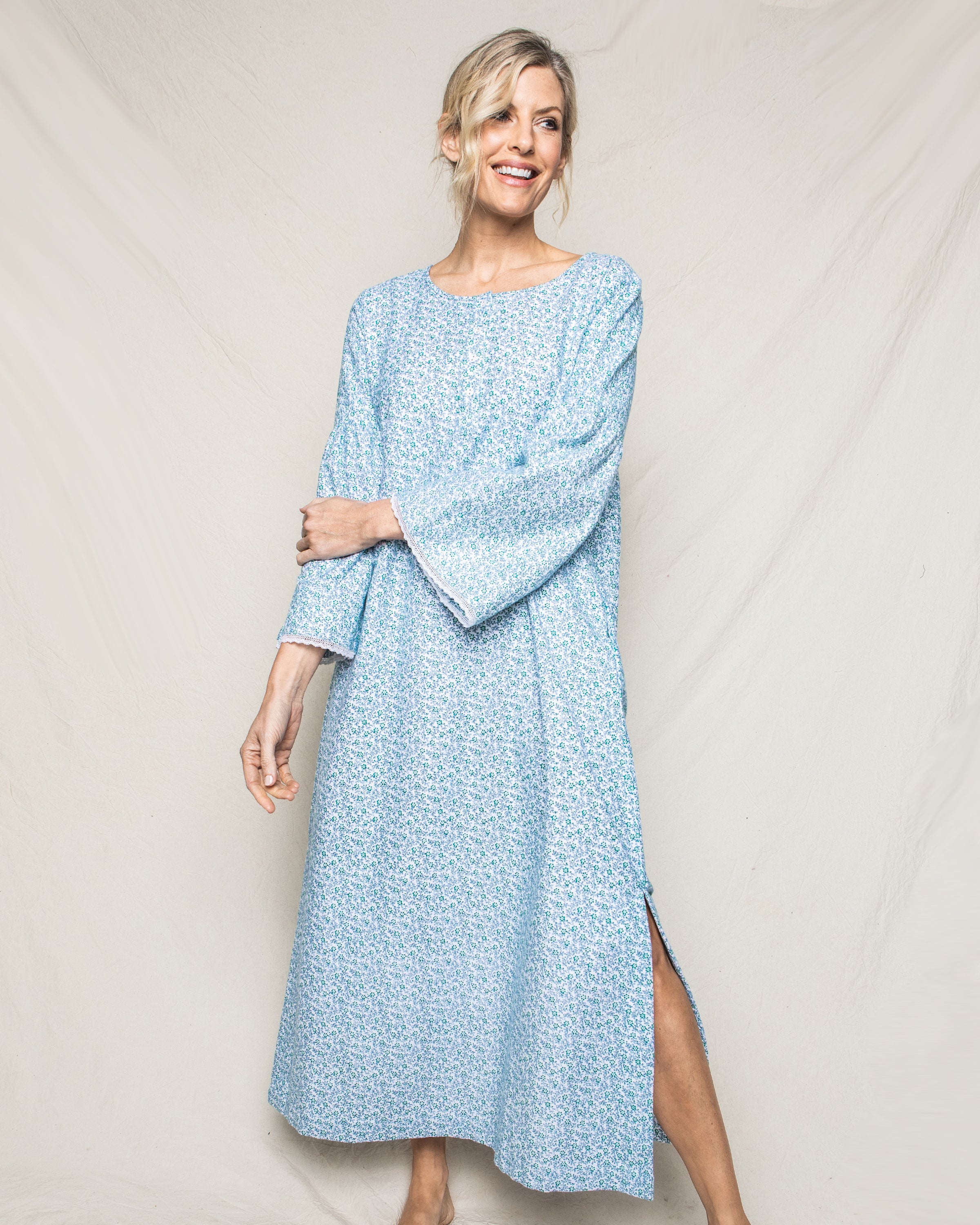 A person wears the Petite Plume Womens Flannel Seraphine Nightgown in Stafford Floral, a long, high-quality cotton garment with subtle patterns and long sleeves plus a side slit. Theyre on a plain backdrop, smiling to the side with one arm bent and the other resting on their hip.