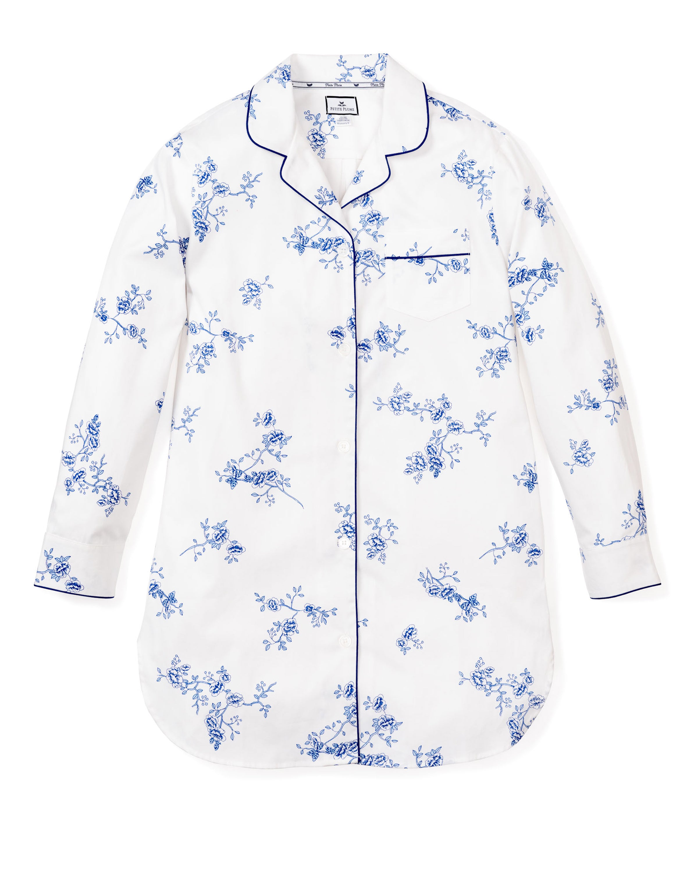 The Womens Twill Nightshirt in Indigo Floral by Petite Plume is a long-sleeved white pajama top adorned with blue floral patterns and blue piping. It features a notched collar and a left chest pocket, combining timeless elegance with cozy comfort.