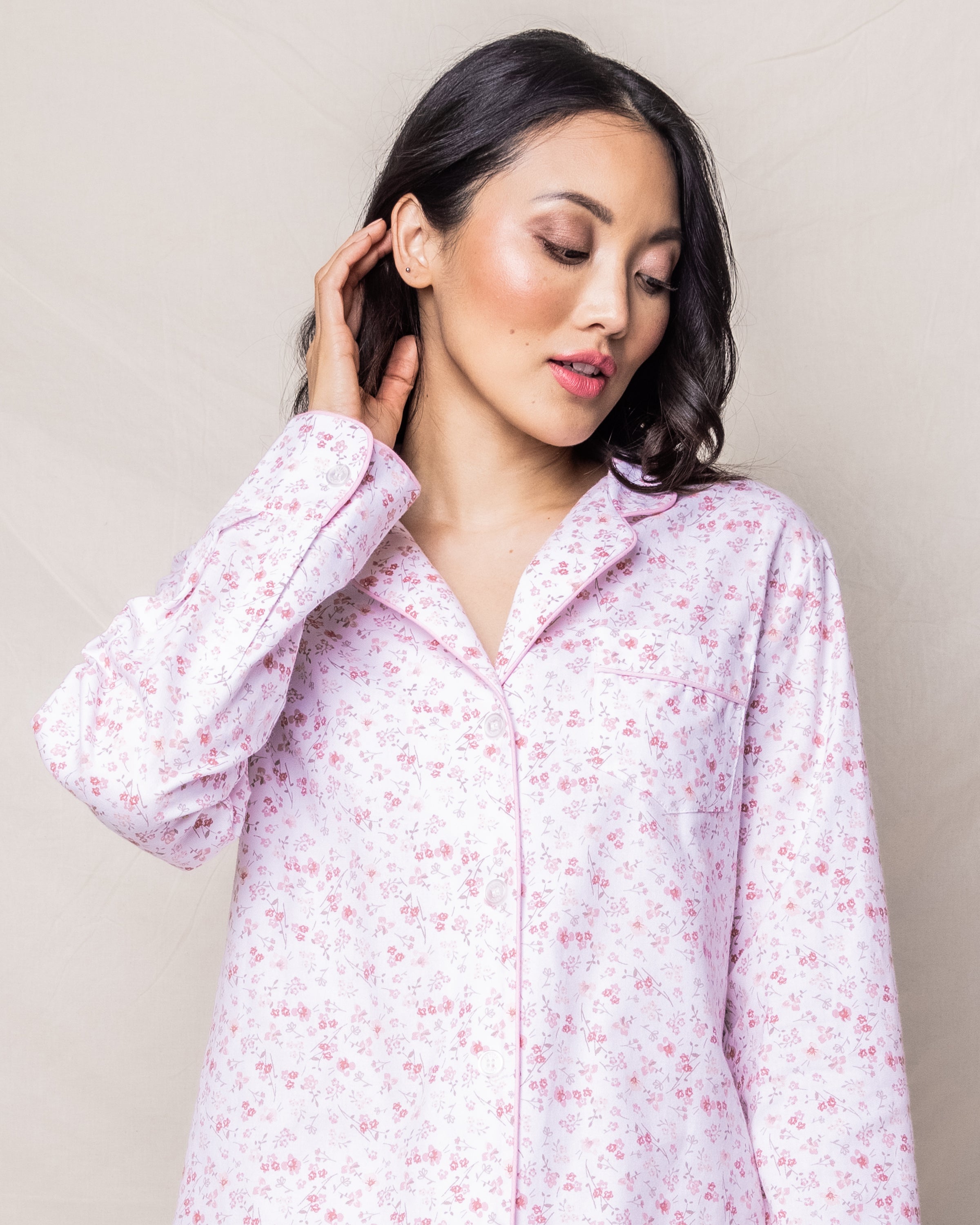 A dark-haired woman elegantly dressed in Petite Plumes Womens Twill Pajama Set in Dorset Floral touches her ear. This luxury sleepwear features a light pink floral design with a collar and pocket, crafted from the finest quality cotton, set against a plain, light background.