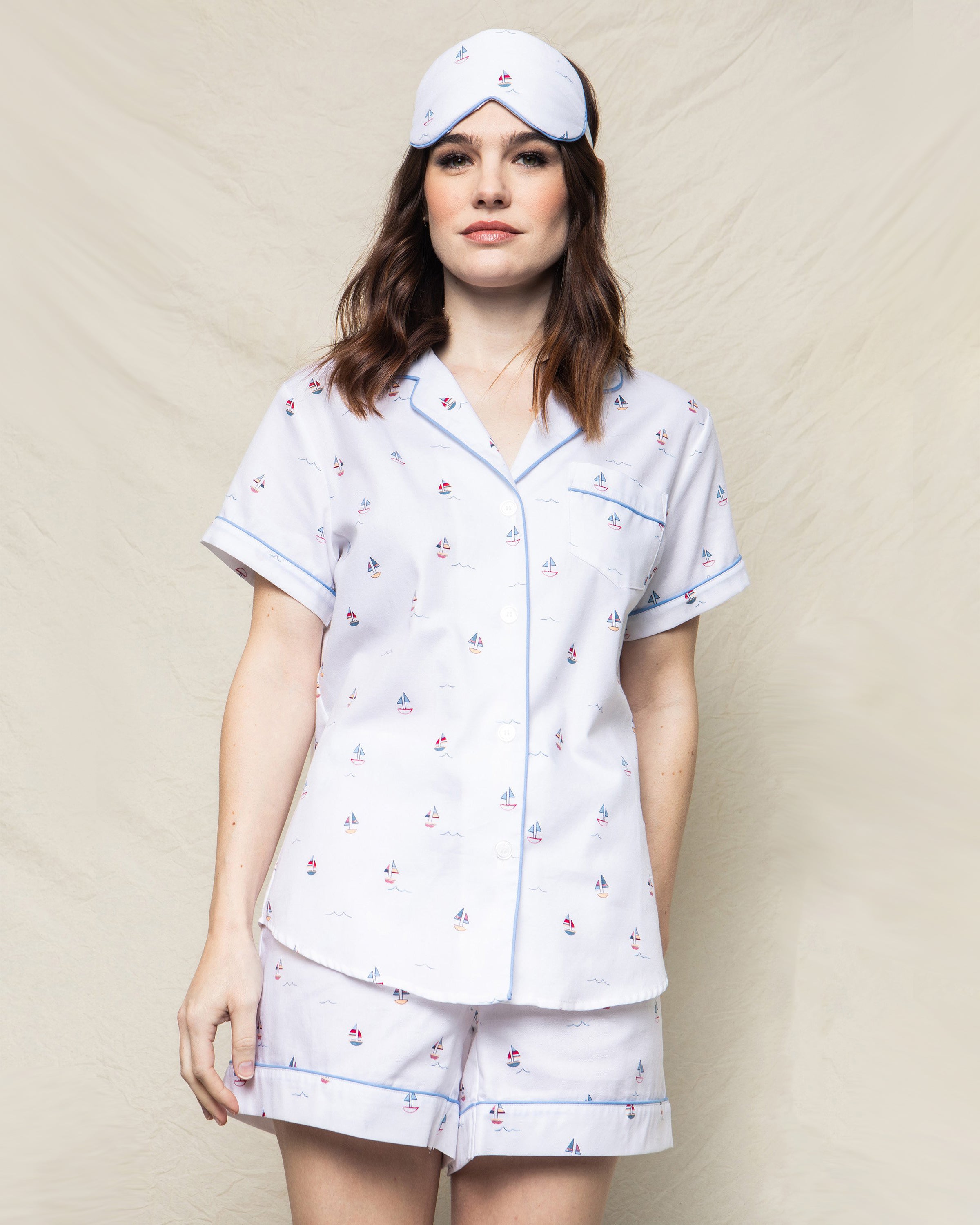 A woman elegantly wears the Womens Twill Short Set in Bateau by Petite Plume, featuring toy boat patterns and light blue trim, paired with a matching sleep mask. She embodies luxury sleepwear against a neutral, textured backdrop.