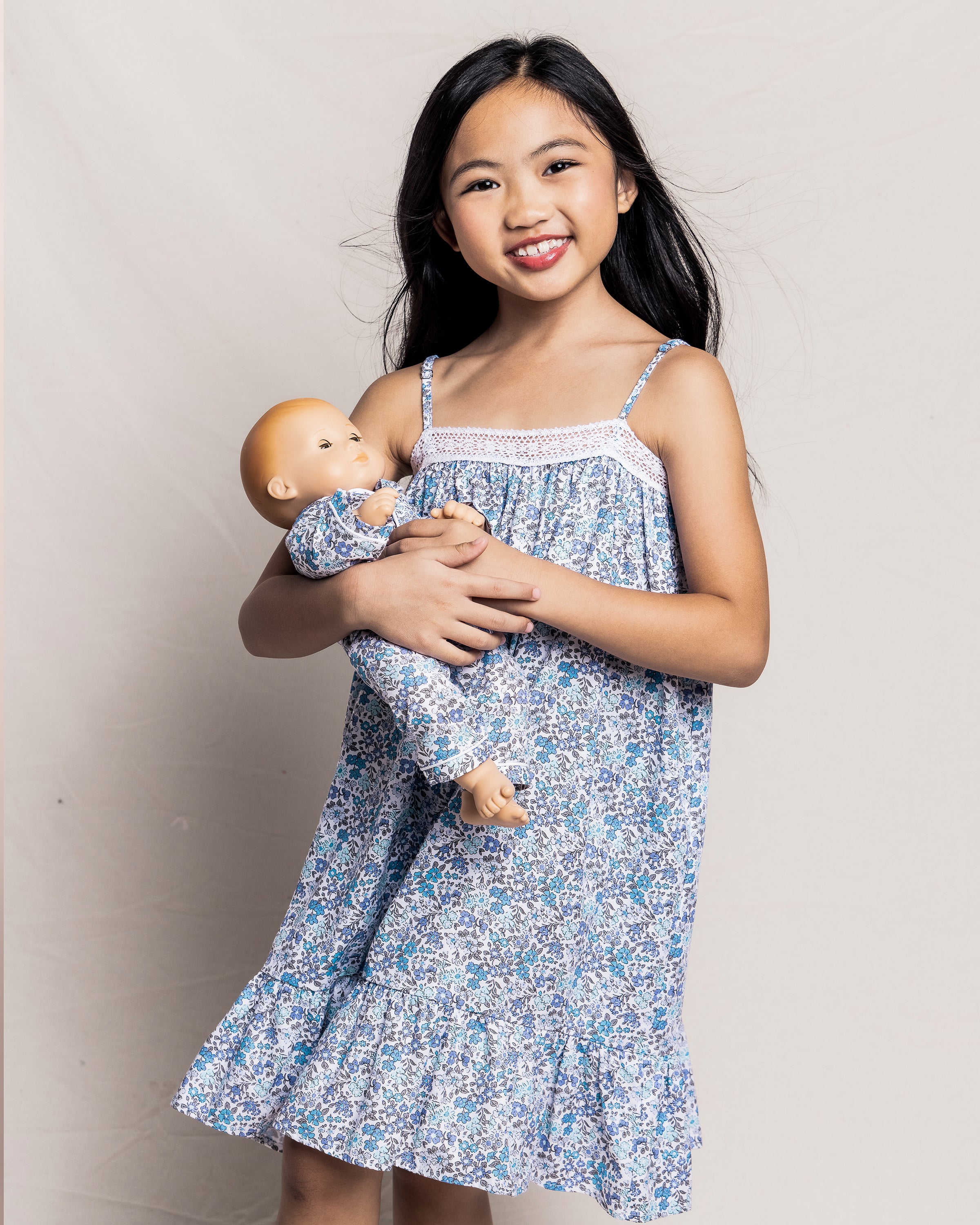 A girl with long black hair, wearing a blue floral dress, smiles and holds an 18 Petite Plume doll in Kids Twill Doll Pajamas Fleur DAzur against a light background.