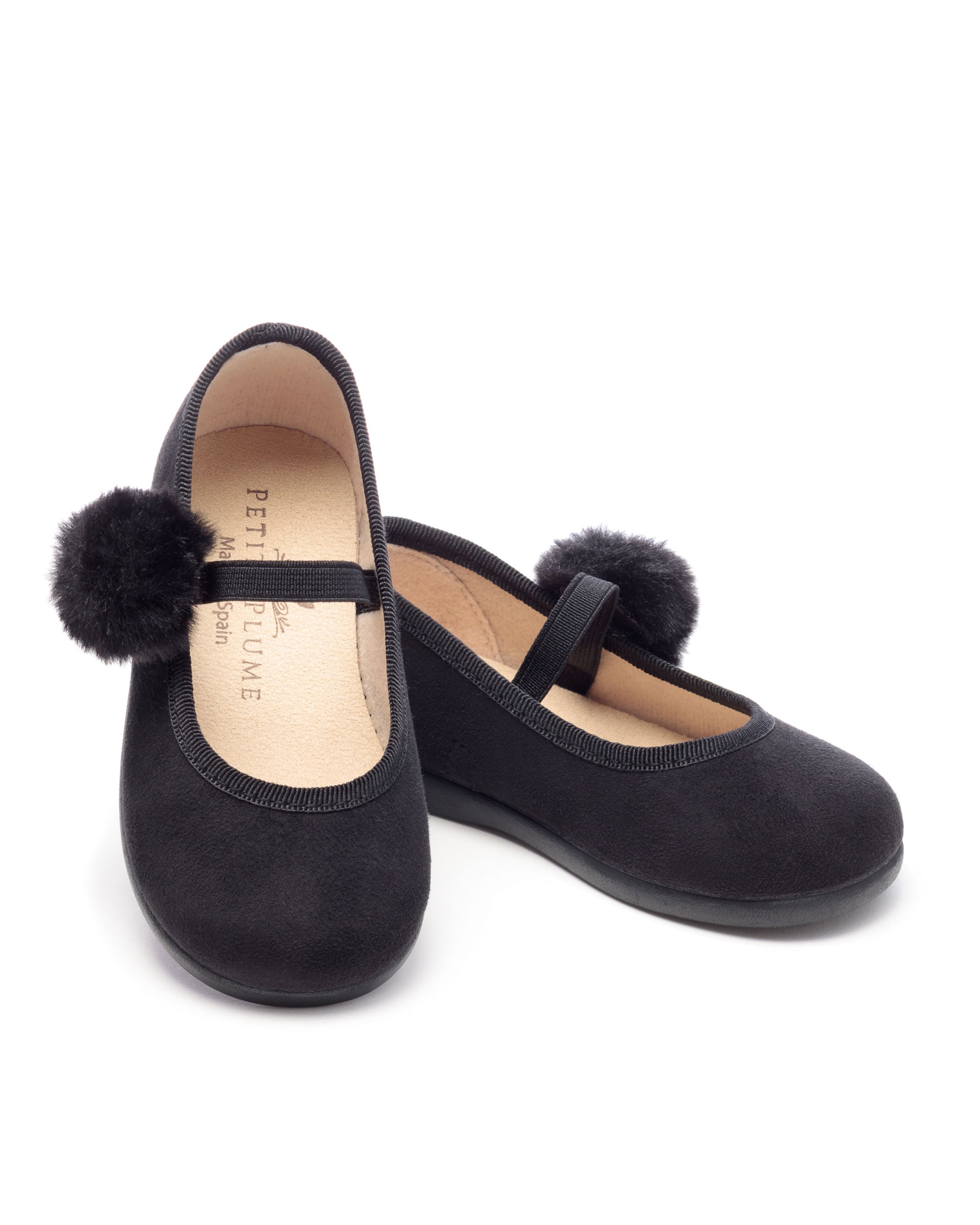 A pair of Kids Delphine Slipper by Petite Plume in black suede displays a festive pom on each toe. One shoe stands upright, while the other leans elegantly against it, showcasing their luxe matte finish and stylish strap design.