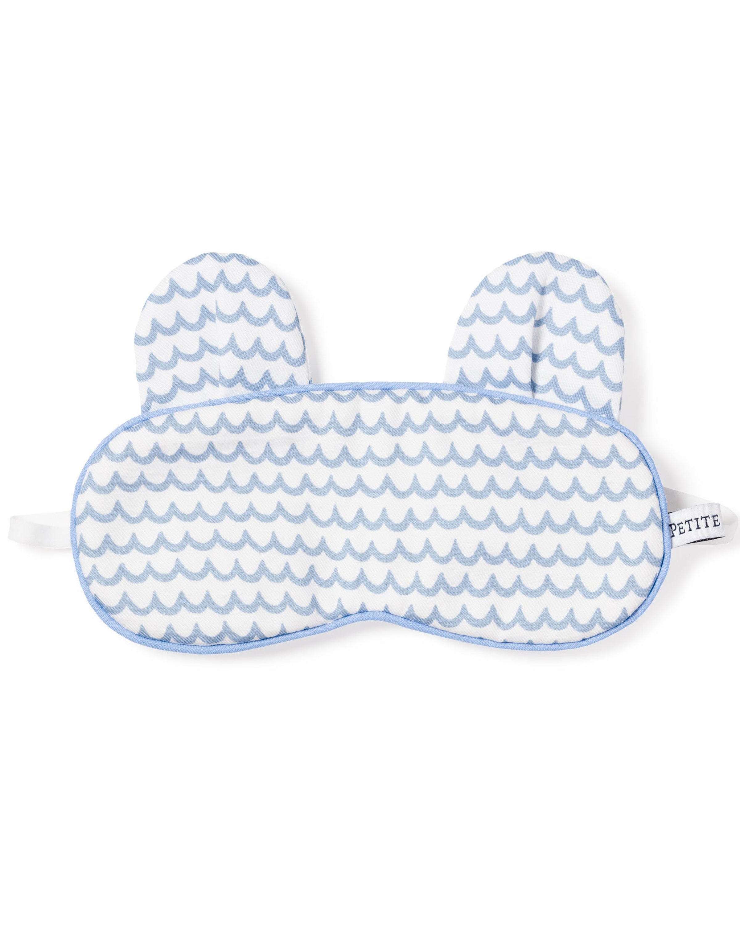 Petite Plumes Kids Bear-y Sleep Mask in La Mer boasts a wavy blue and white design, ear-like extensions, and a sleek white strap. A monogrammed tag on the right adds elegance, perfect for stylish kids who love matching eye masks.