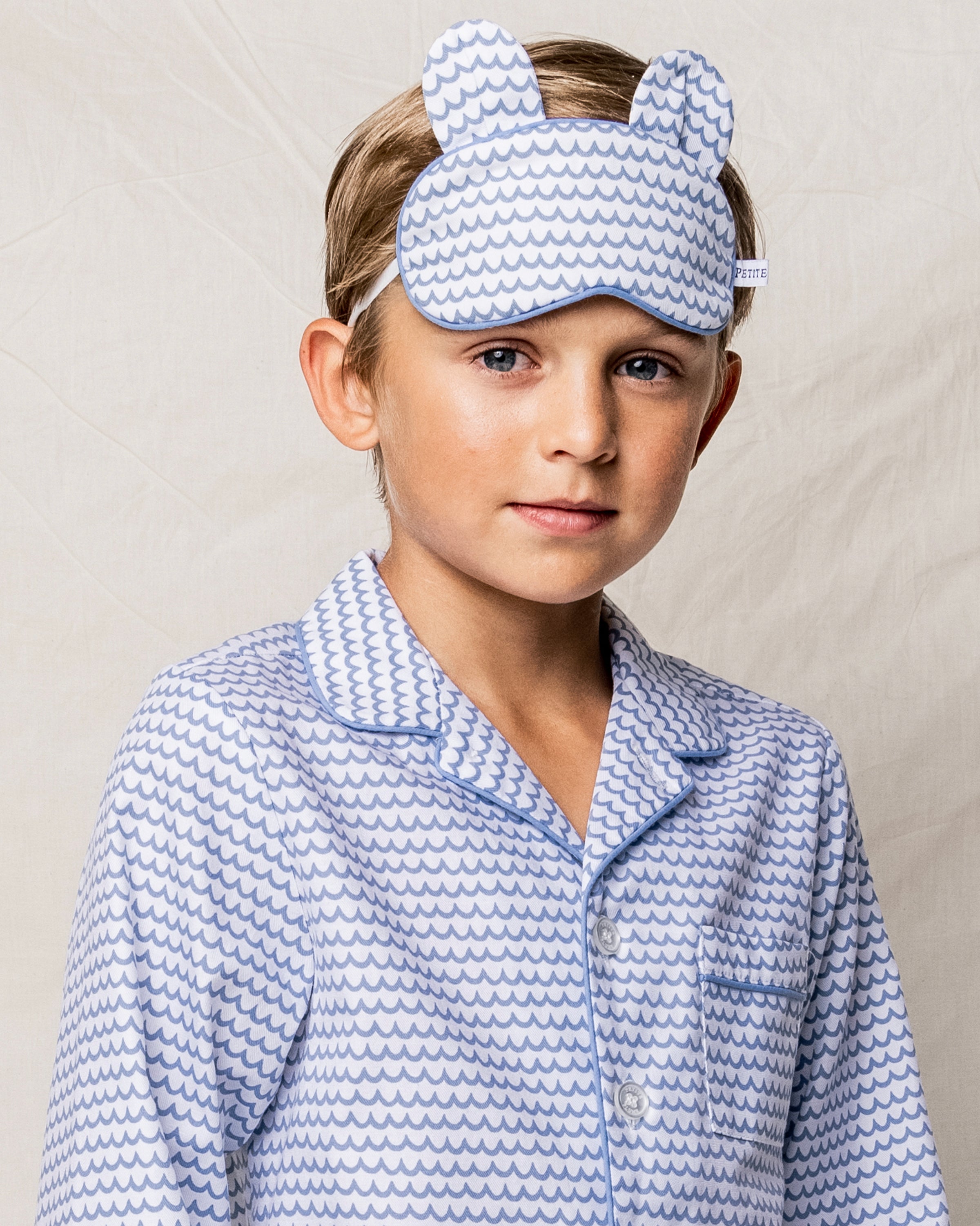 A child in patterned blue and white pajamas enjoys luxury with the matching Petite Plume Kids Bear-y Sleep Mask in La Mer, featuring cute small animal ears. The plain light fabric background enhances the cozy elegance.