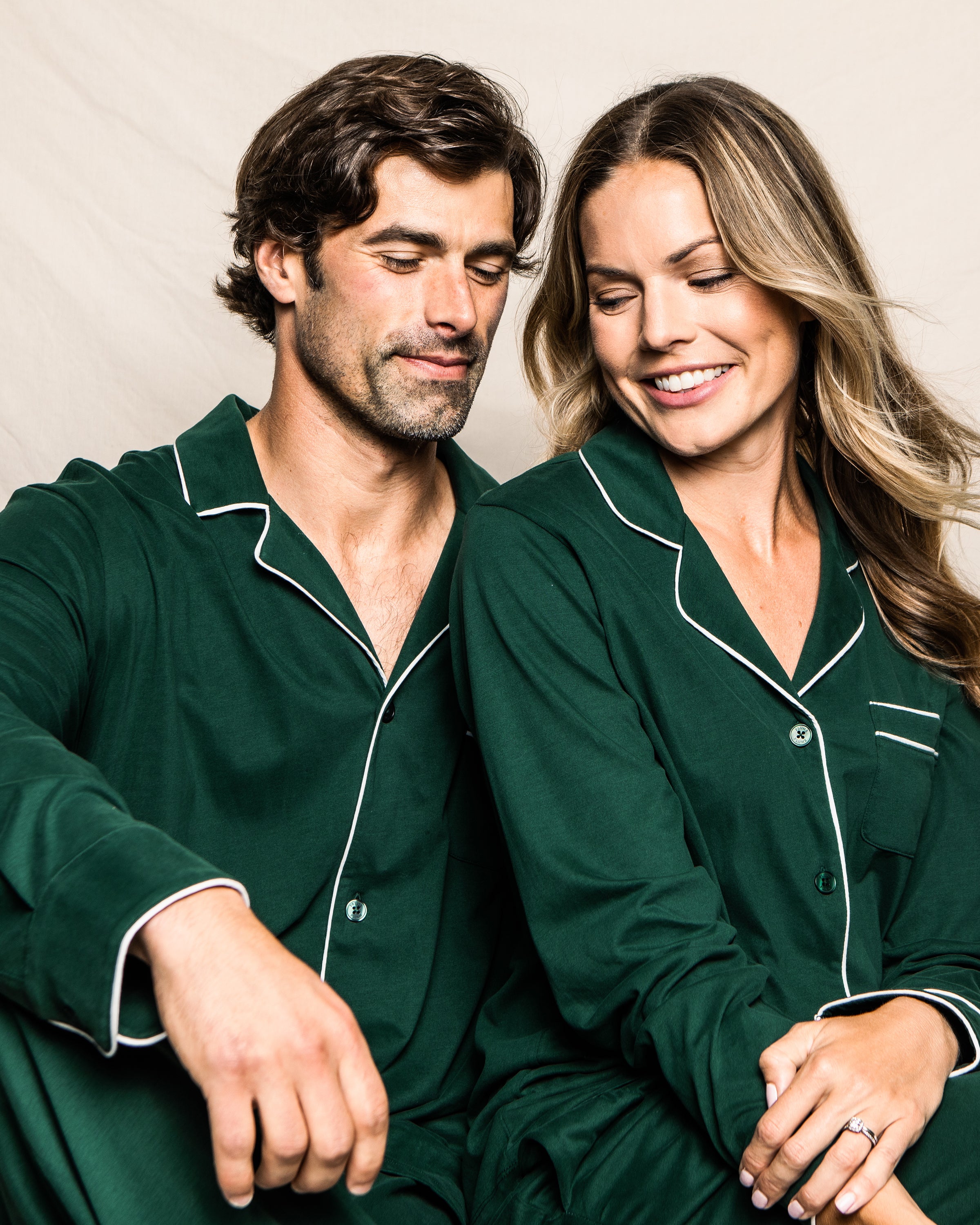 A couple in matching Petite Plumes Mens Pima Pajama Set in Evergreen sits against a neutral backdrop, smiling. The womans gaze shifts downward while the mans hand rests on his knee, and hers hovers close by, illustrating elegance and timeless comfort.