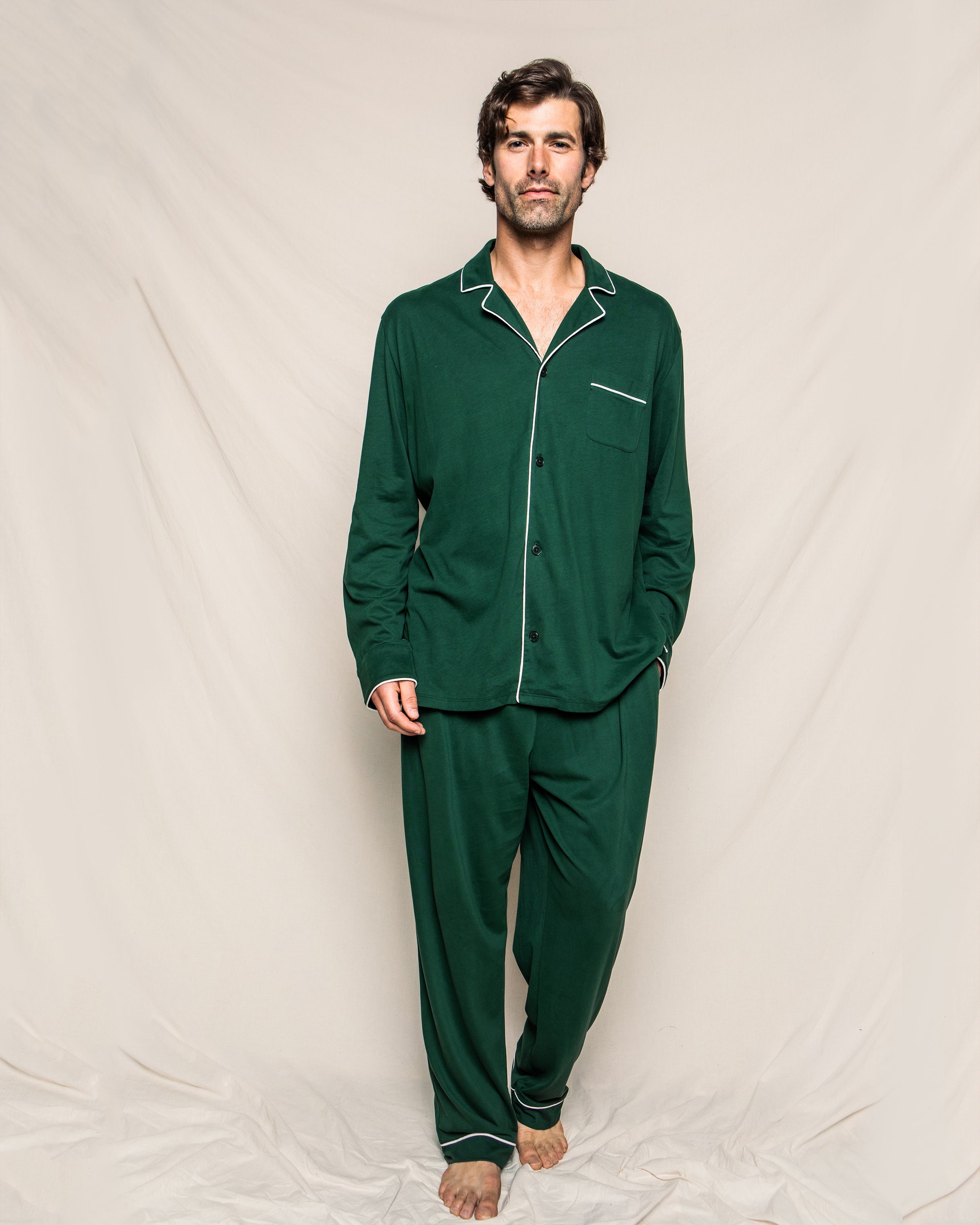 Wearing the Mens Pima Pajama Set in Evergreen by Petite Plume, a person poses against a neutral backdrop. This luxurious Peruvian Pima cotton set features classic white trim, a collared button-up top, and matching pants for a timeless, relaxed, and comfortable look.