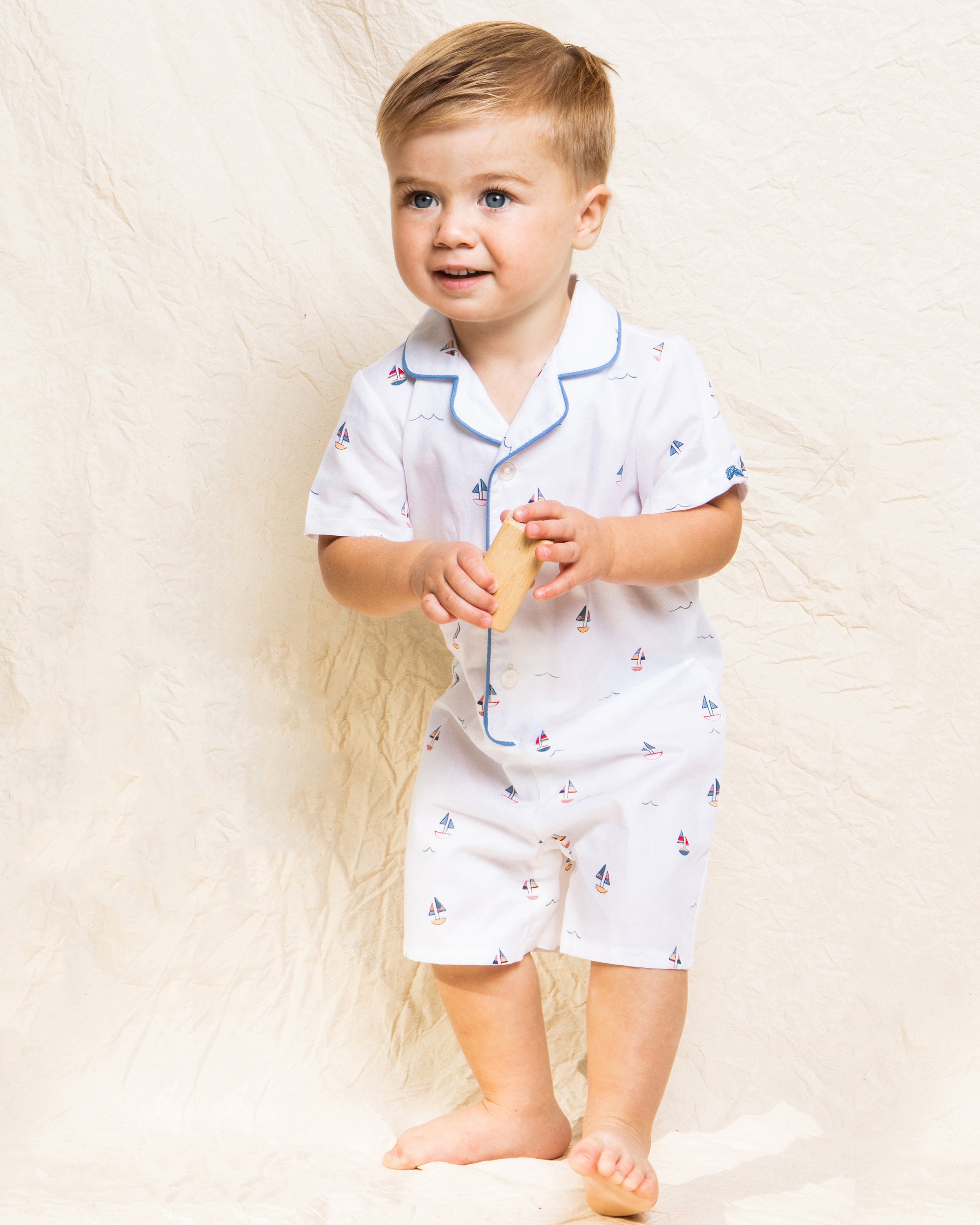 A toddler wearing Petite Plumes Babys Twill Summer Romper in Bateau stands barefoot on a light fabric background, holding a wooden toy and smiling slightly to the side.