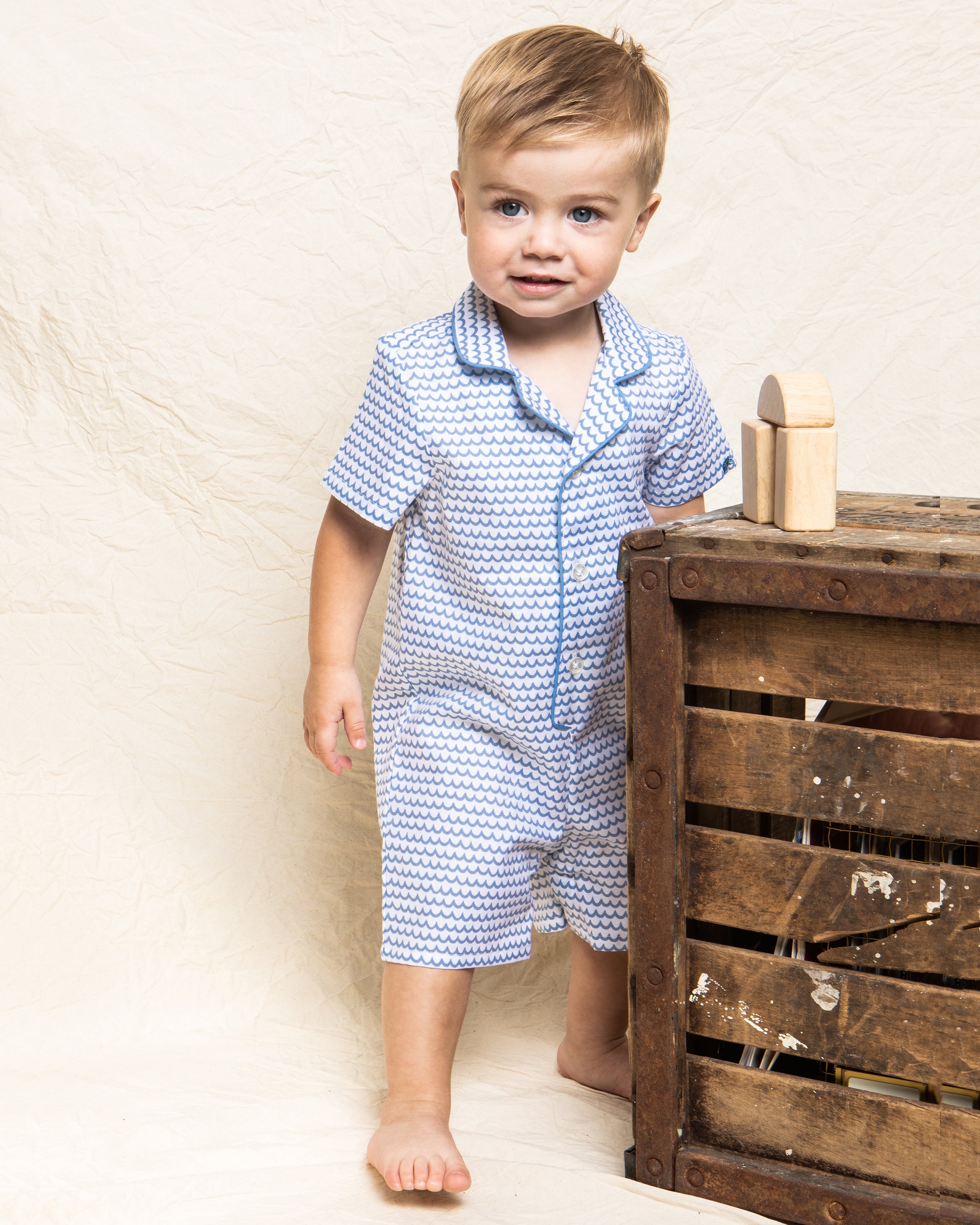 A toddler in Petite Plumes Babys Twill Summer Romper in La Mer stands barefoot by a rustic wooden crate, smiling softly with a building block on the crate. The background features textured, off-white fabric.