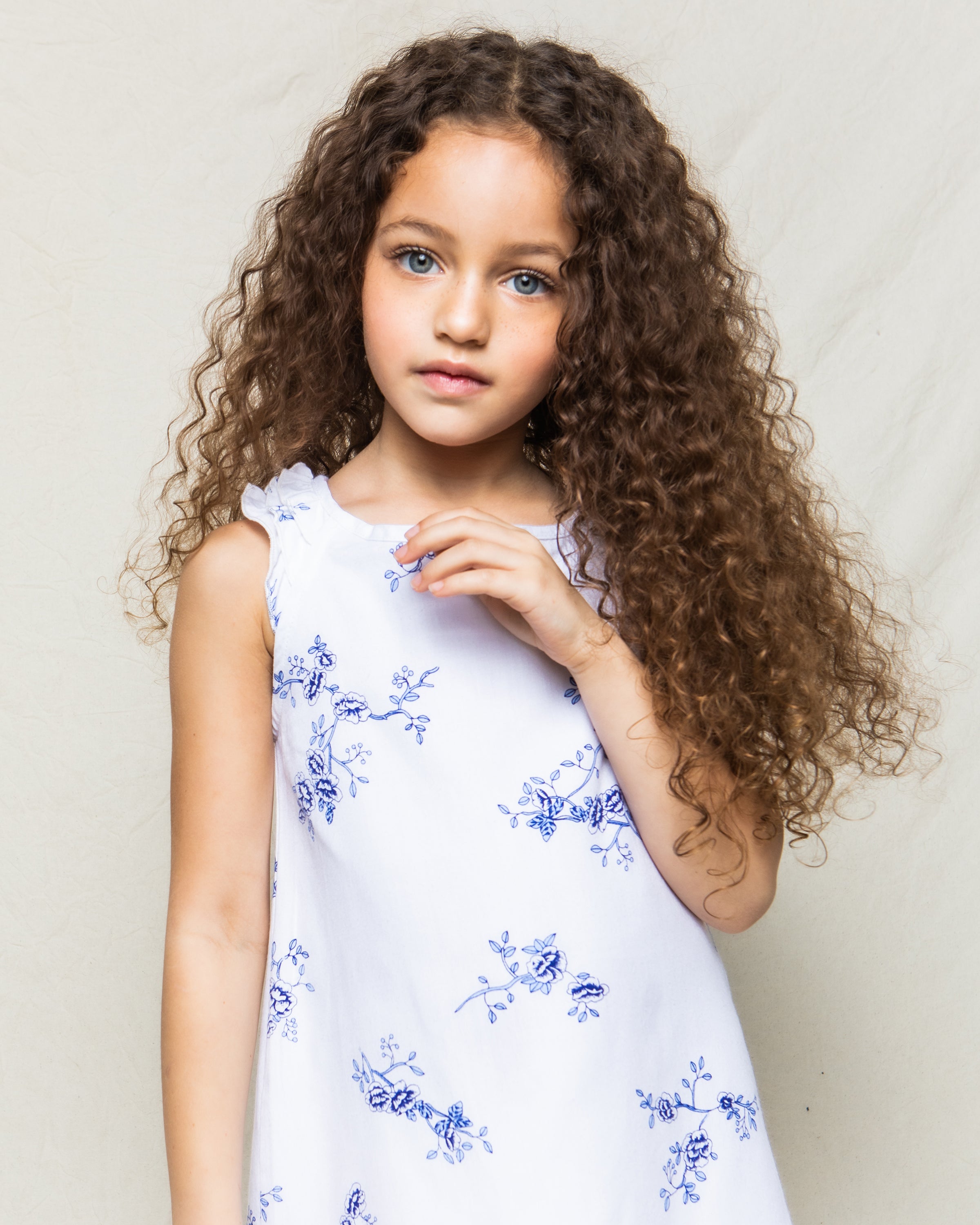 A young girl with long, curly hair wears the Petite Plume Girls Twill Amelie Nightgown in Indigo Floral. Crafted from high-quality cotton, she stands against a light, neutral background, gazing calmly at the camera.