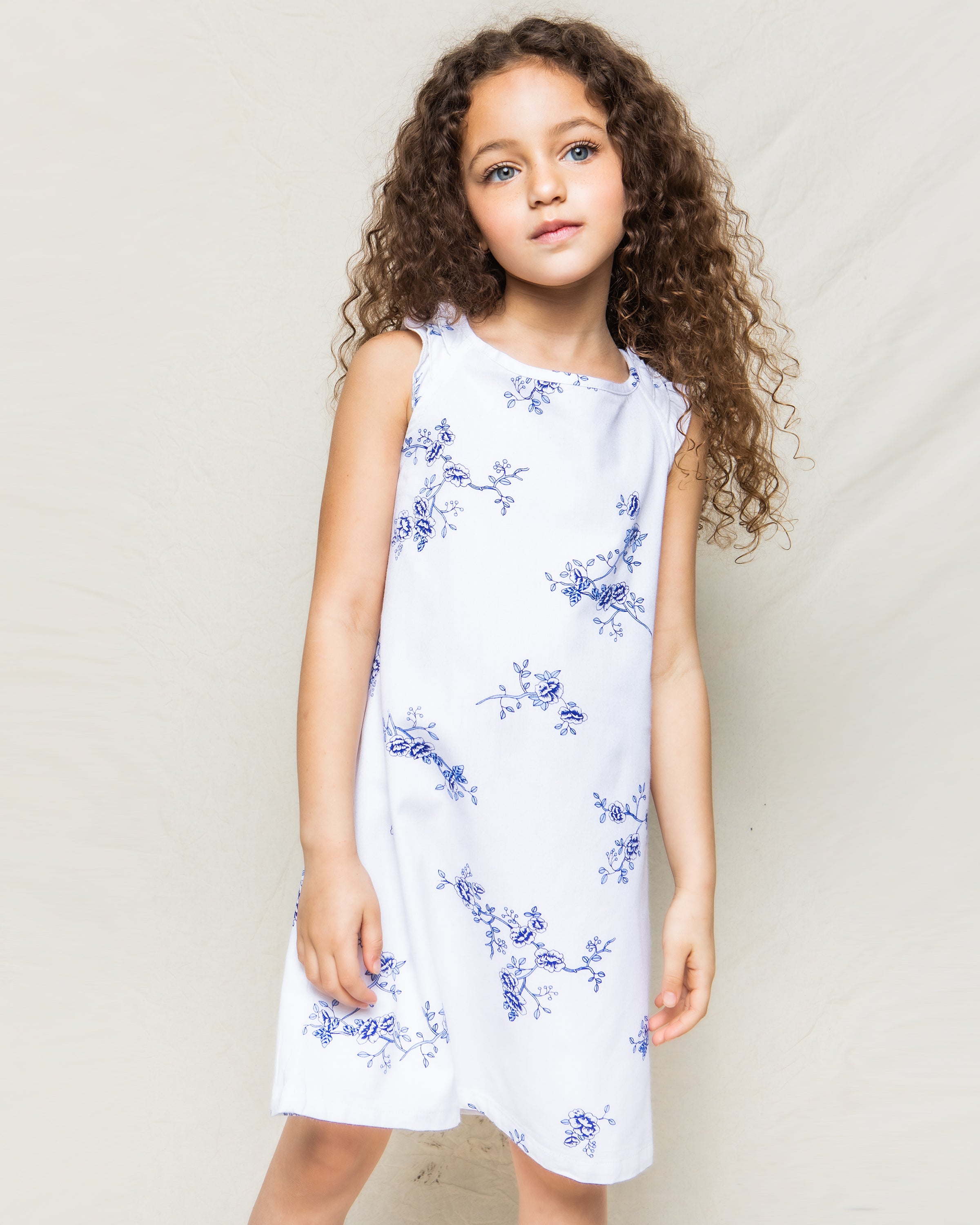 A young girl with long curly hair wears Petite Plumes sleeveless Girls Twill Amelie Nightgown in Indigo Floral, crafted from the finest cotton, standing against a plain light background.