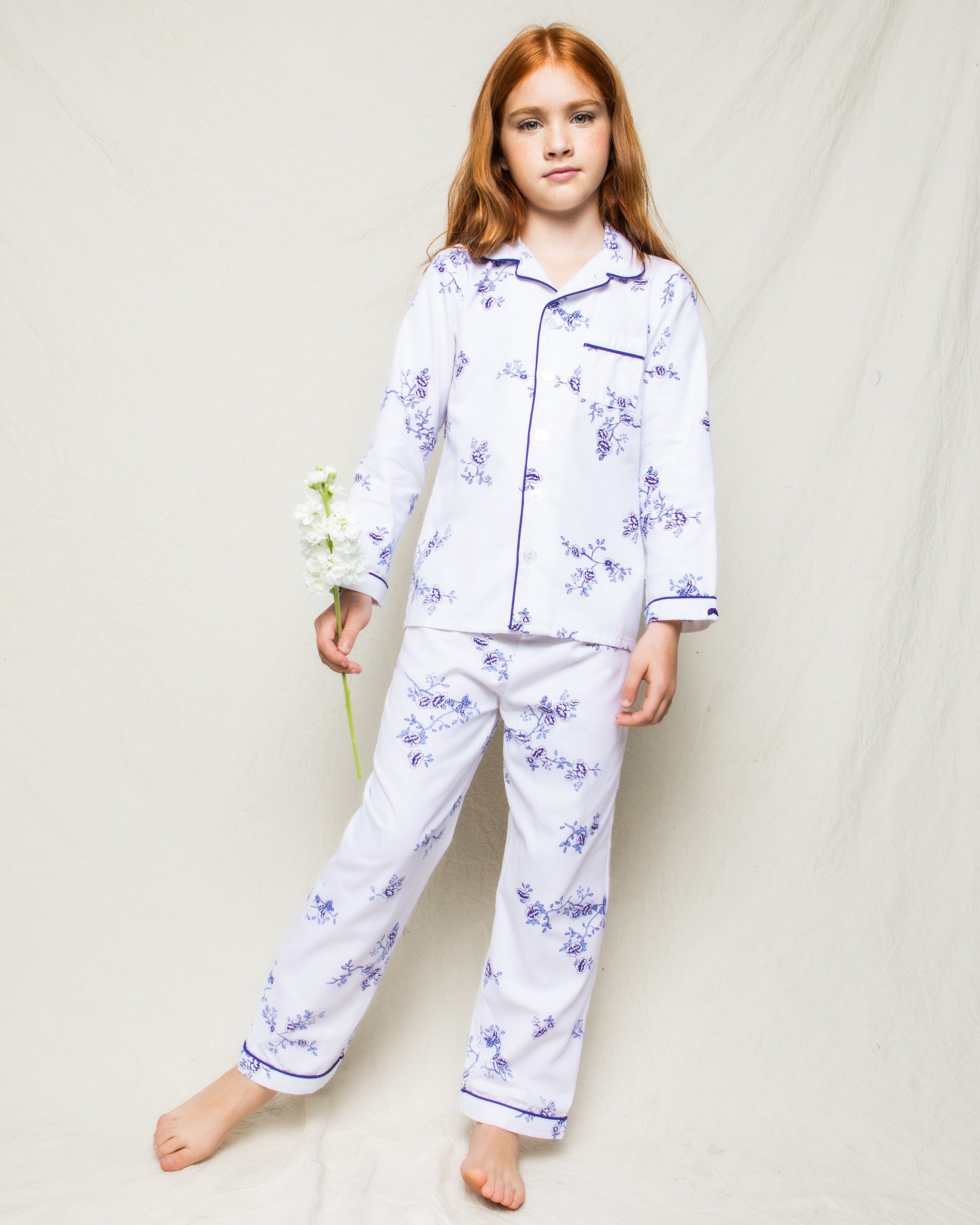 A young girl with long red hair stands barefoot against a plain background, wearing Petite Plumes Kids Twill Pajama Set in Indigo Floral. She holds a small bouquet of white flowers in her right hand, exuding an air of serene elegance.