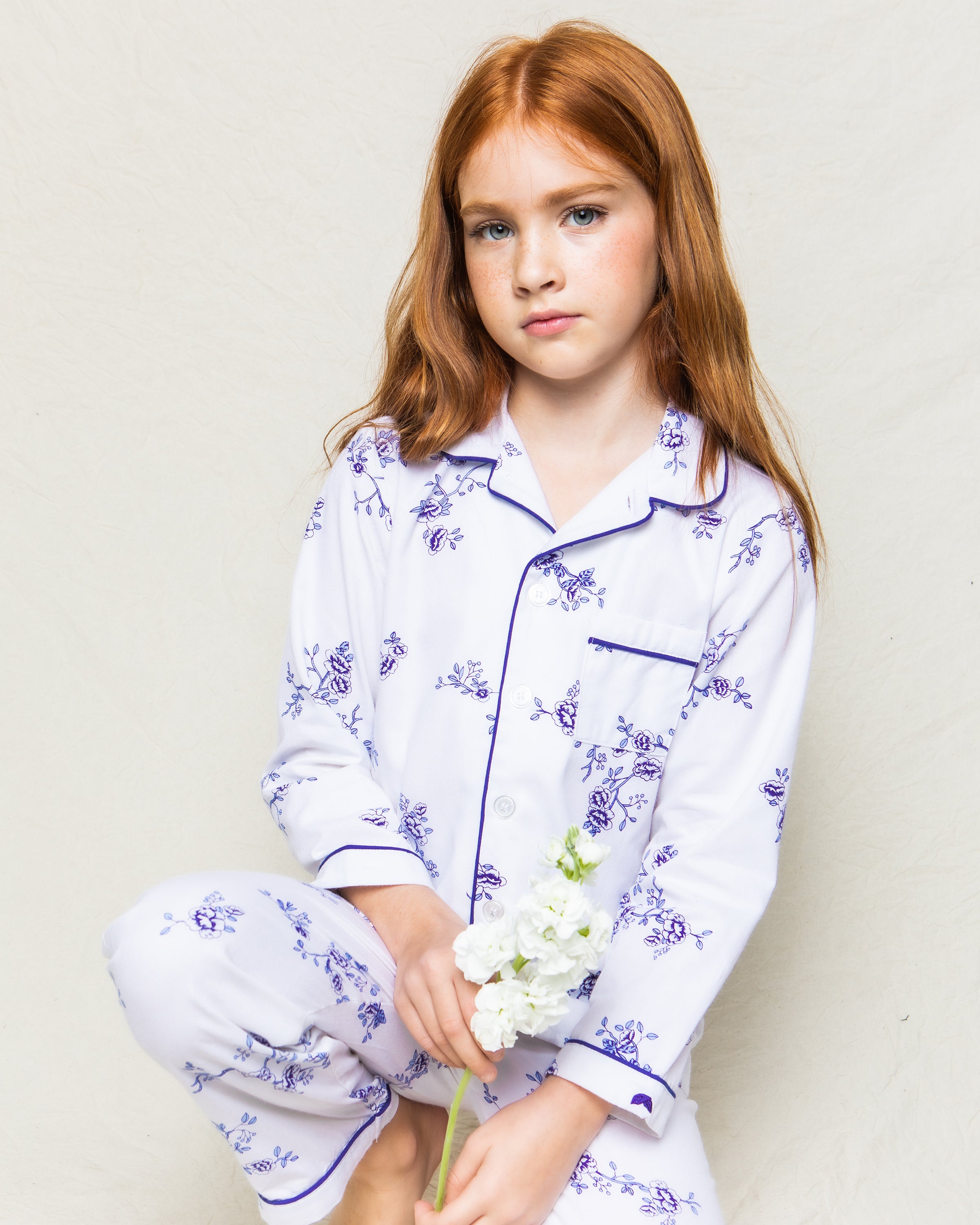 A young girl with long red hair wears the Kids Twill Pajama Set in Indigo Floral by Petite Plume. She holds a white flower and looks at the camera neutrally, set against a light gray or cream background, adding vintage charm.