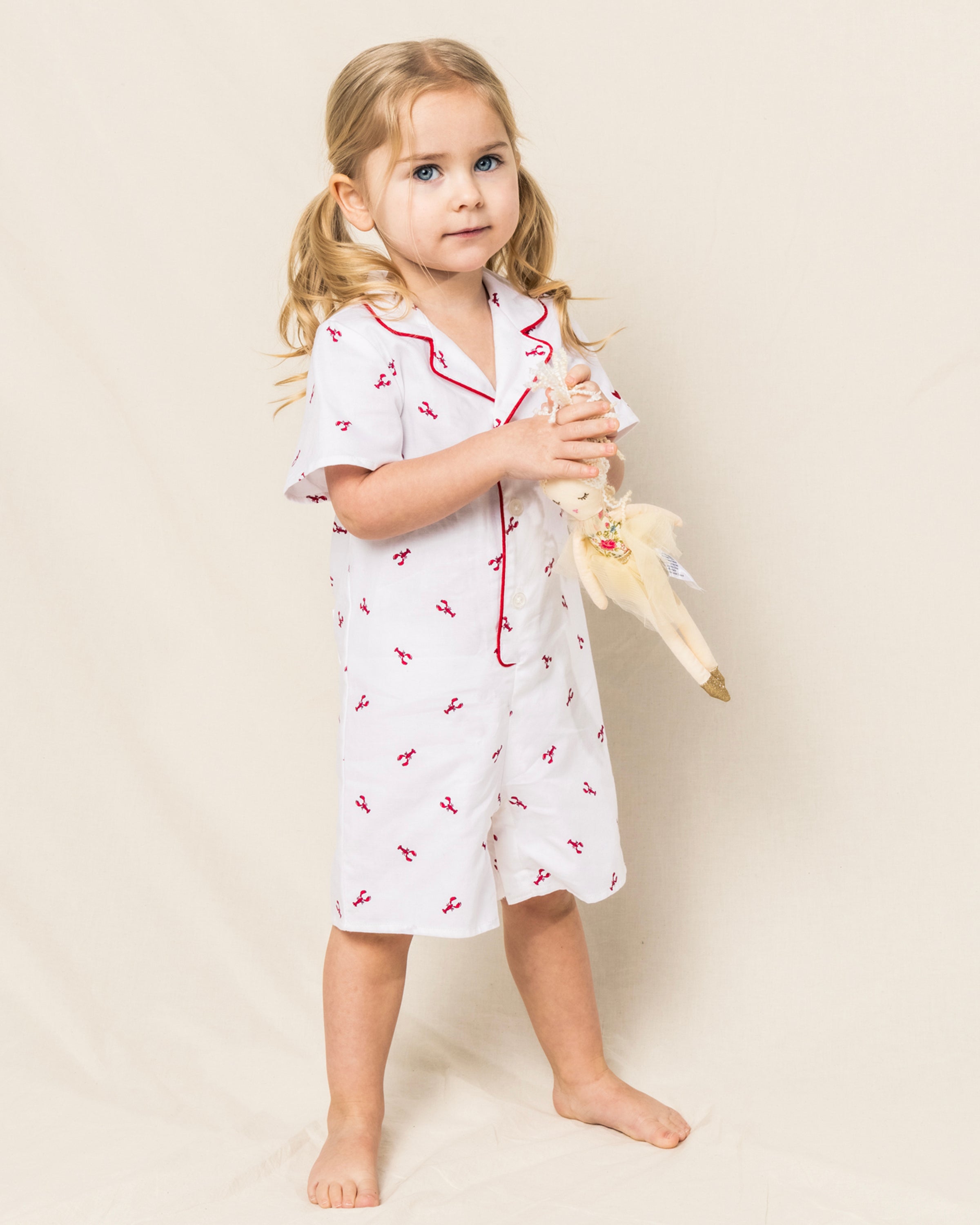 A young blonde child in pigtails wears Petite Plumes Baby Twill Summer Romper in Brixham Lobsters, crafted from soft brushed fabric. The white romper has red designs, piping, and short sleeves. She stands barefoot against a light-colored background holding a doll.