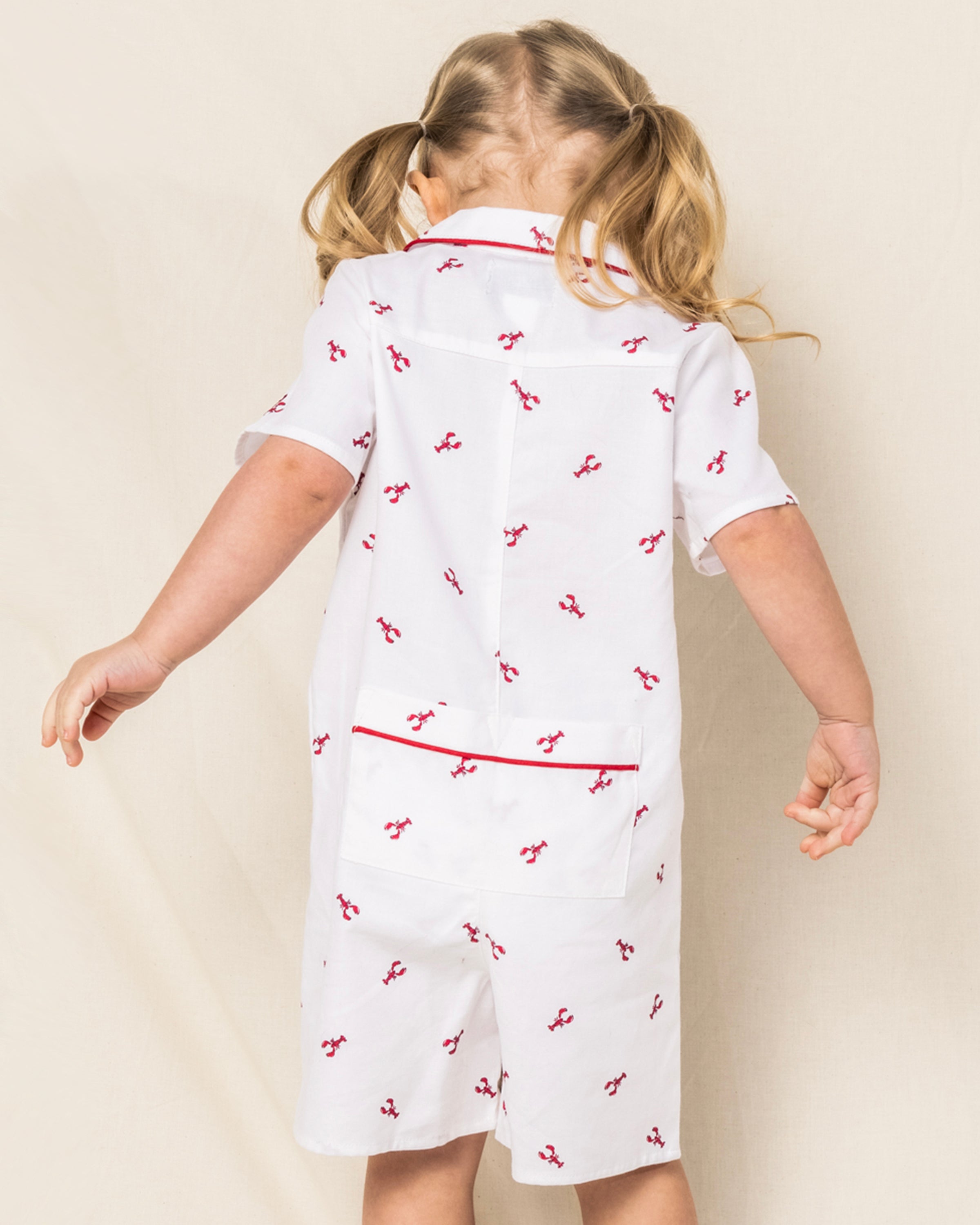 A child with blonde pigtails, facing away, wears Petite Plumes Babys Twill Summer Romper in Brixham Lobsters. Crafted from the softest brushed fabric, it features small red cherry patterns and a back pocket, with red trim on the white short sleeves against a neutral beige background.