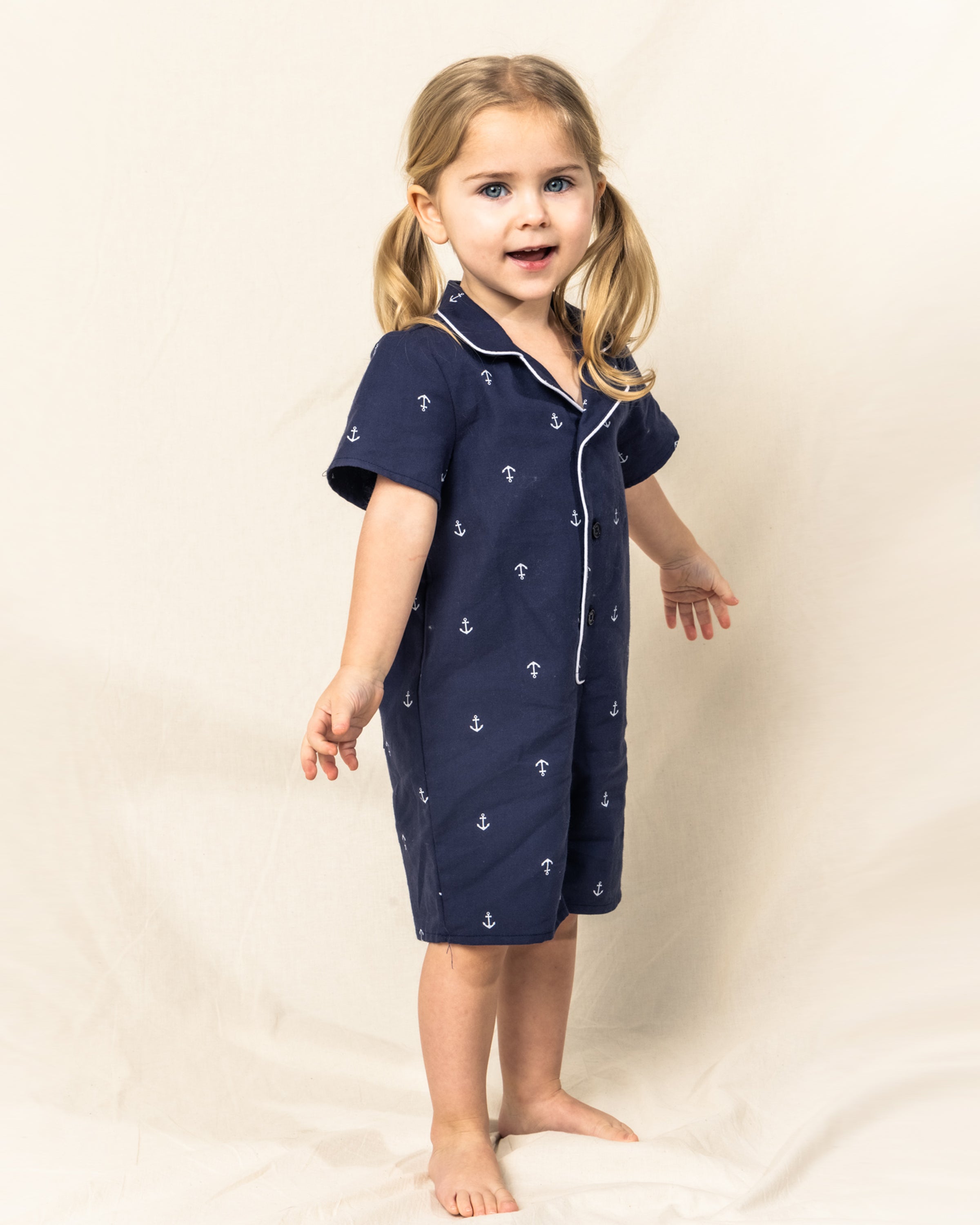 A young child with blonde pigtails stands barefoot on a cream background, wearing Petite Plumes Babys Twill Summer Romper in Portsmouth Anchors. The navy blue romper has an anchor pattern with white piping. Her arms are slightly outstretched as she gazes forward curiously.