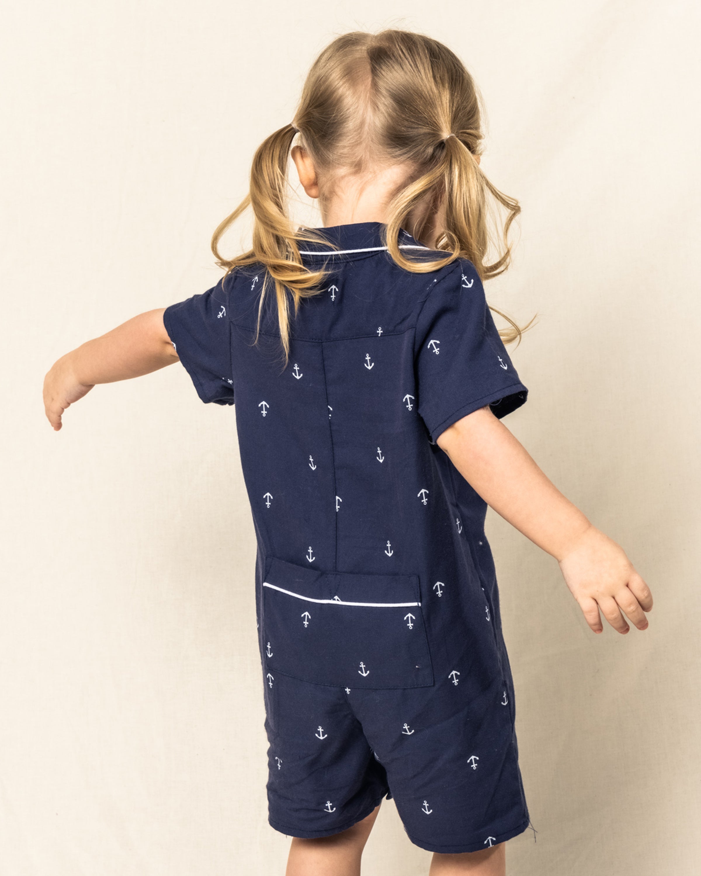 A young child with blonde pigtails, facing away, wears the Petite Plume Babys Twill Summer Romper in Portsmouth Anchors. This navy romper is adorned with white navy anchors and crafted from yarn-dyed cotton for comfort and style against a plain beige background.