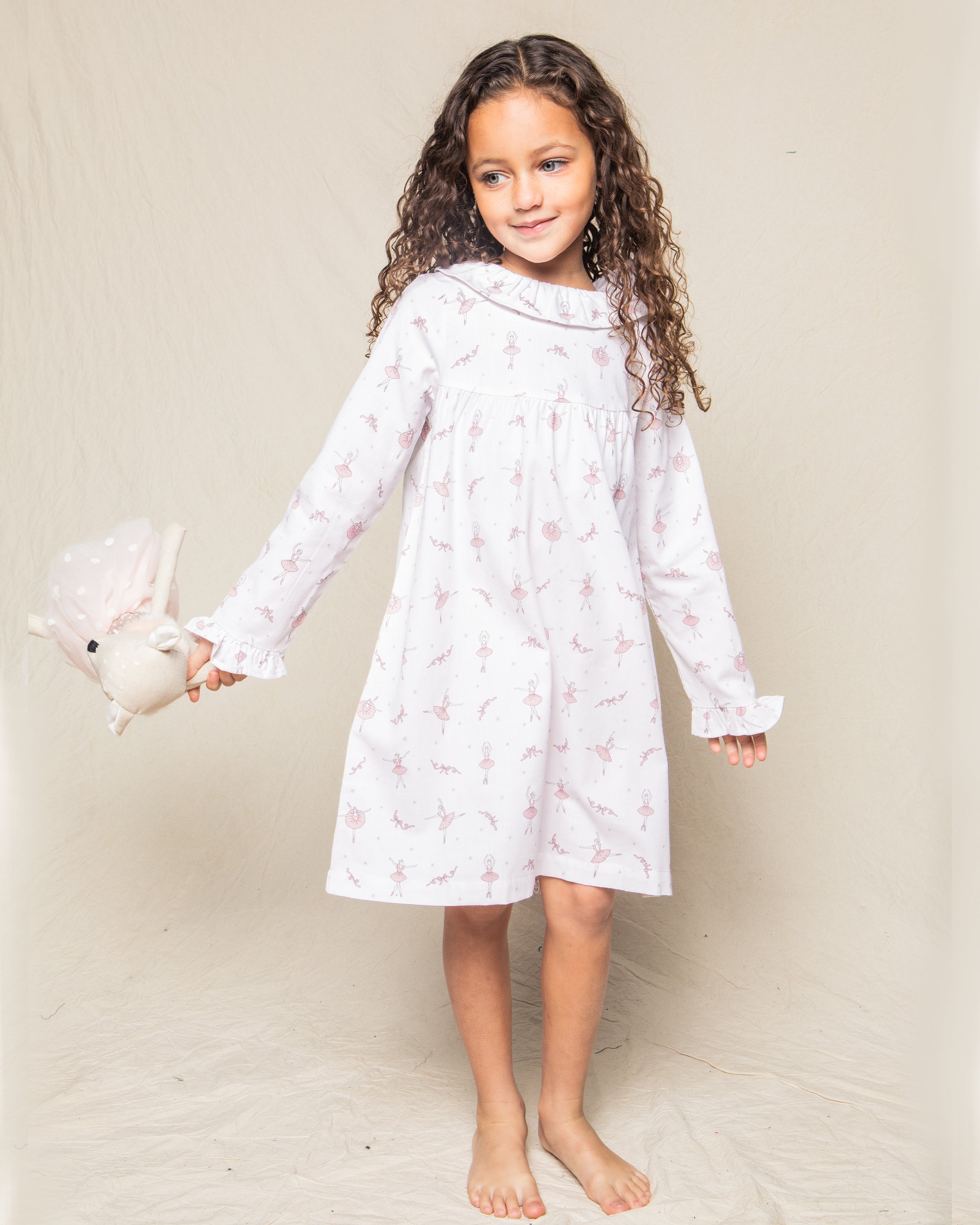 A young girl with long curly hair stands barefoot, wearing a Petite Plume Girls Twill Scarlett Nightgown in Sugar Plum Fairy. She holds a stuffed bunny toy and smiles gently against a neutral background.