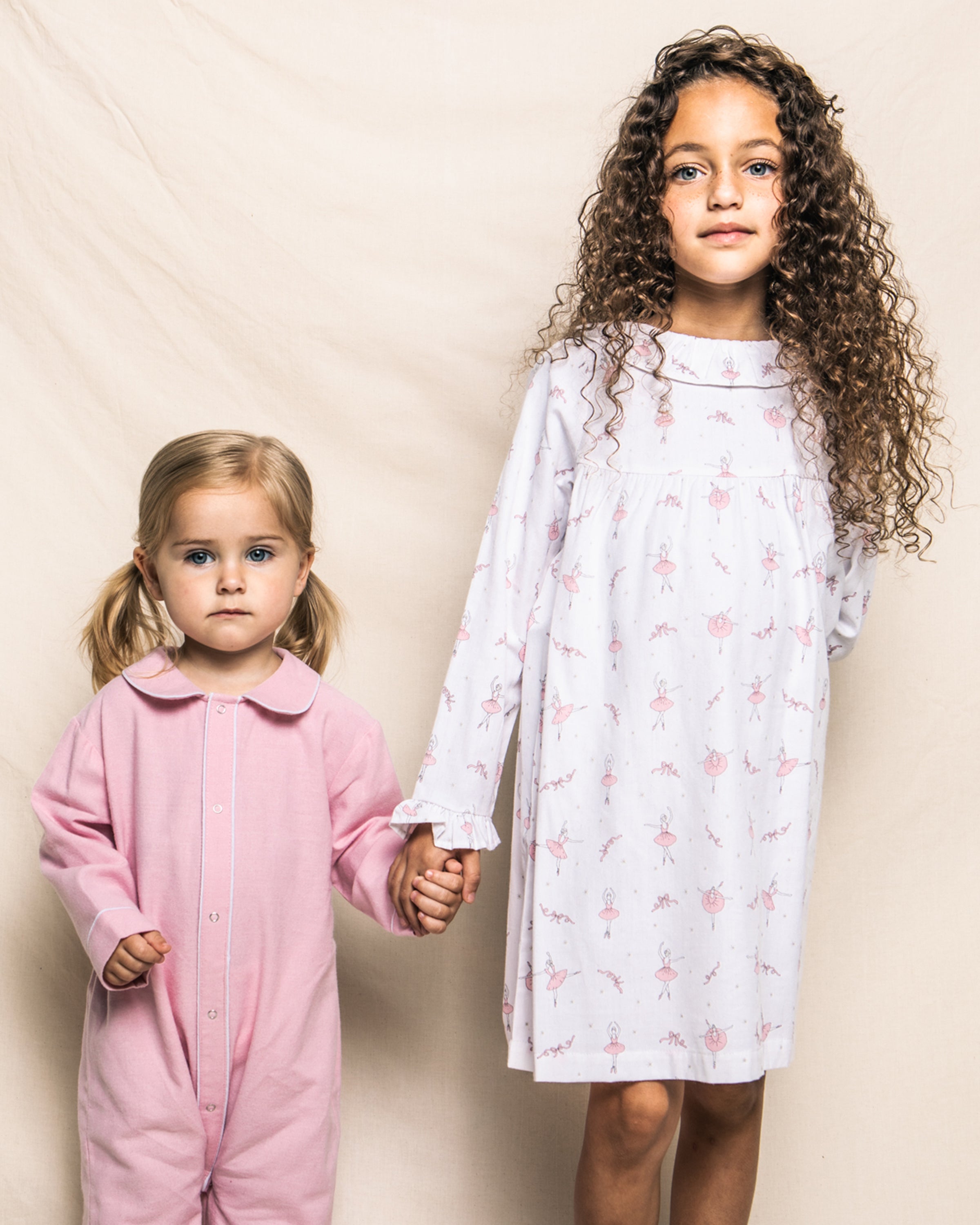 Two young children stand before a light backdrop. The younger child on the left wears a pink onesie with pigtails. The older child, with long curly hair, gently holds hands with them while wearing Petite Plumes Girls Twill Scarlett Nightgown in Sugar Plum Fairy, made from flame-retardant fabric.